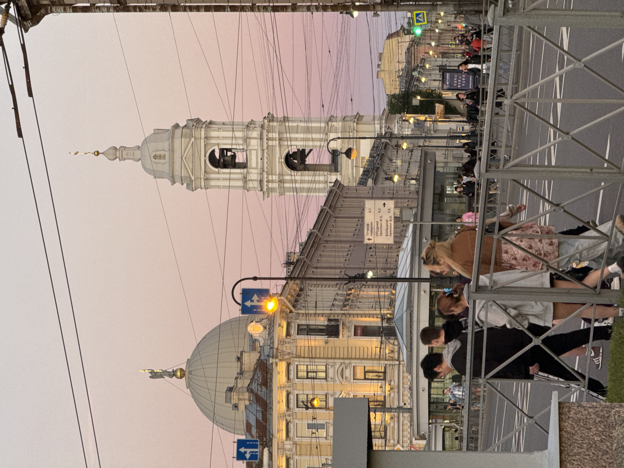 This is a medium-shot, eye-level photograph of a street scene in Saint Petersburg, Russia, during what appears to be twilight. In the foreground, a group of four young people are walking across a street, partially obscured by a grey metal railing. The person closest to the viewer is a woman with blonde hair, wearing a brown jacket and a floral print dress. To her left is a person wearing a grey hoodie and dark shorts, followed by a young man in a black hoodie and black pants, and another young man in a grey hoodie and dark pants. The street itself is paved with asphalt and marked with white lines.
In the middle ground, a wide street with multiple lanes of traffic is visible. There are buildings lining the street on both sides, featuring classical architecture with ornate detailing. A prominent element is a tall, white bell tower with a clock face and arched openings containing bells. To the left of the bell tower is a large, light-colored dome topped with a golden statue of an angel. Streetlights are illuminated, casting a warm glow. Several traffic signs and electrical wires are visible throughout the scene. Pedestrians can be seen walking on sidewalks and crossing the street in the distance.
The background features a soft, pinkish-purple sky, indicating either dawn or dusk. The overall atmosphere is calm and urban. The buildings suggest a historic district within Saint Petersburg, likely near a significant landmark given the architectural style and the presence of a large dome and bell tower. The presence of streetcars or trams is implied by the overhead wires.