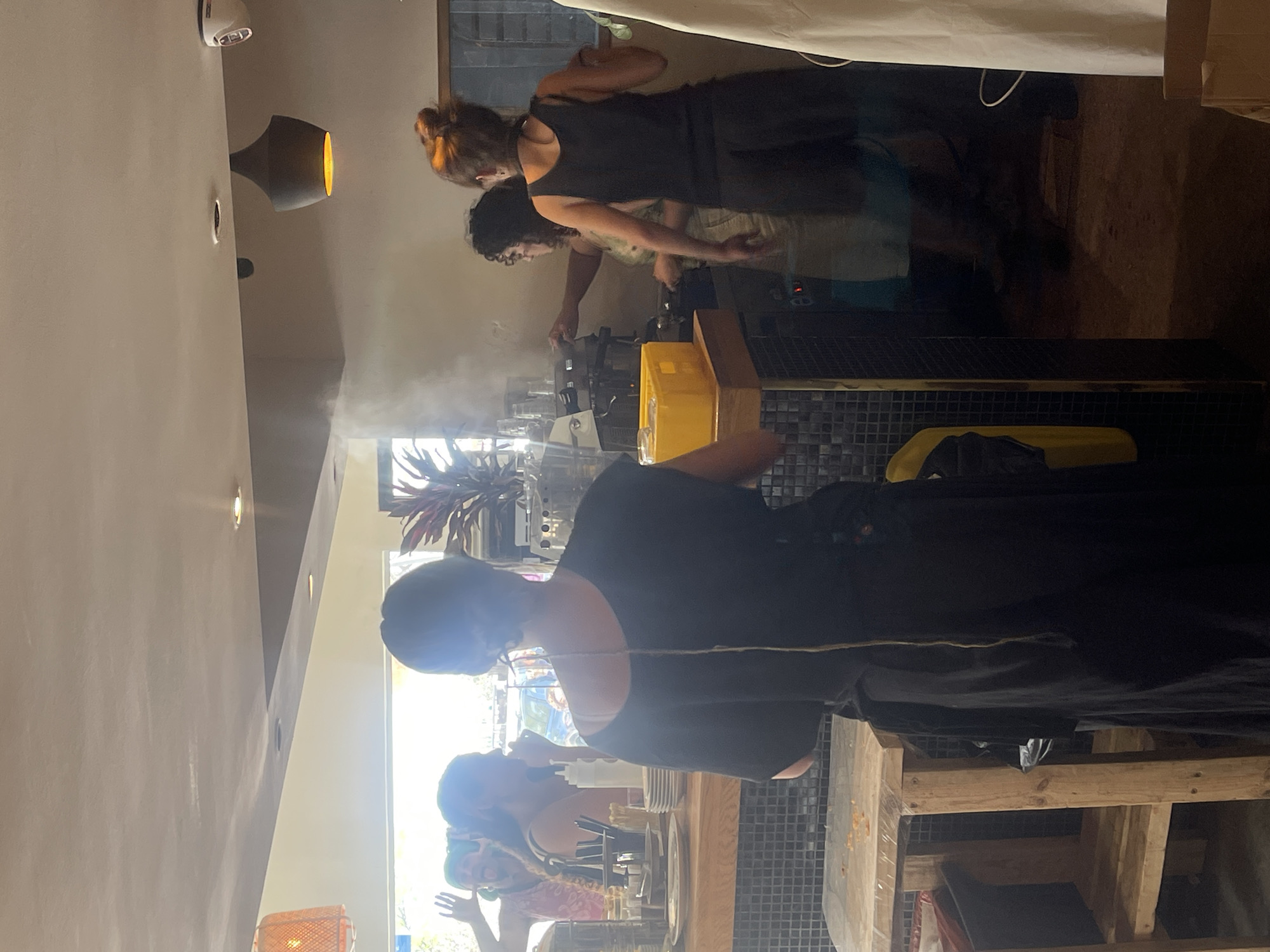 The original image, rotated 90 degrees clockwise, depicts an active indoor scene, likely a cafe or restaurant kitchen/serving area, during daytime in Mitzpe Ramon, Israel. At least five individuals are visible, engaged in various tasks.
In the mid-ground, behind a black tiled counter, two people appear to be working. One person with reddish-brown hair, tied back, is operating an espresso machine or similar equipment, which is prominently emitting a large cloud of steam or smoke, suggesting the preparation of hot beverages. Another individual with dark, possibly curly hair, and wearing a lighter top, stands beside them, also behind the counter. In the foreground, a person with dark, short hair and a black top stands with their back mostly to the camera, a thin cord or string visible across their back. Further back and to the right, a person with green hair can be seen gesturing or looking towards others. An arm and shoulder of another person are visible near a wooden structure in the lower right.
The setting features modern, slightly rustic elements including a black tiled counter with a metallic trim, wooden shelving, and warm-toned ceiling lights; one is a black, bell-shaped fixture with an orange glow. Bright natural light streams in from large windows, illuminating the space, complemented by artificial lighting. Yellow crates or containers are visible on the counter, alongside various bottles, glasses, and kitchen utensils. Decorative plants, including one with red leaves, add to the ambiance. The atmosphere appears busy and functional. No discernible text is visible in the image.