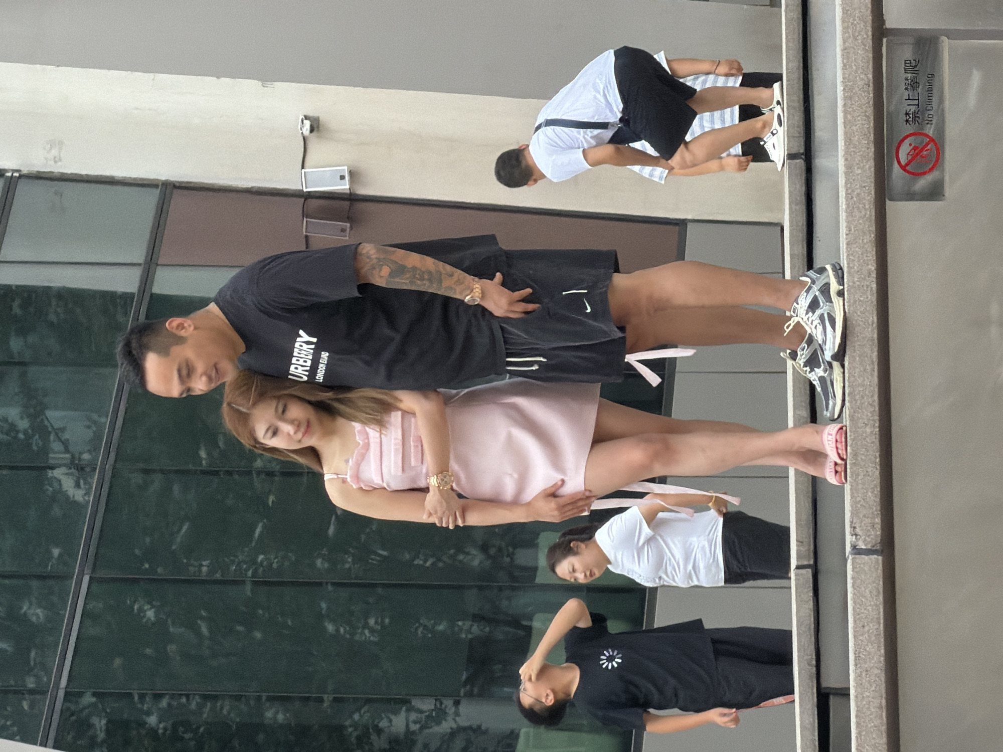 This is a full-shot, eye-level photograph taken outdoors in Yuzhong District, China. The scene depicts a group of people on a multi-tiered stone structure, likely a public space with a modern building in the background.
In the foreground, a man and a woman are standing close together. The man, on the right, is wearing a black t-shirt with "BURBERRY LONDON EST." printed on it in white, and black shorts. He has tattoos on his right arm. The woman, on the left, is wearing a light pink sleeveless dress. Her head is resting on the man's shoulder, and her left hand is holding his right arm. Both are wearing athletic footwear.
Behind them, to the left, a young boy in a black t-shirt and black pants is looking upwards and appears to be touching his face. Further back, a woman in a white t-shirt and black pants is standing and looking to her left.
On the right side of the image, another man in a white t-shirt and black shorts is bent over, possibly interacting with a person or object out of frame. Two other individuals, possibly children, are partially visible behind him, wearing striped tops and shorts.
The background features a large building with a facade composed of dark green glass panels and lighter colored sections. There are electrical conduits and two small white speaker-like devices mounted on the wall of the building. A metal sign with a red circle and diagonal line over an icon of a person climbing, along with Chinese characters and "No Climbing" in English, is visible on the lower right section of the stone structure. The lighting suggests it is daytime, possibly overcast.