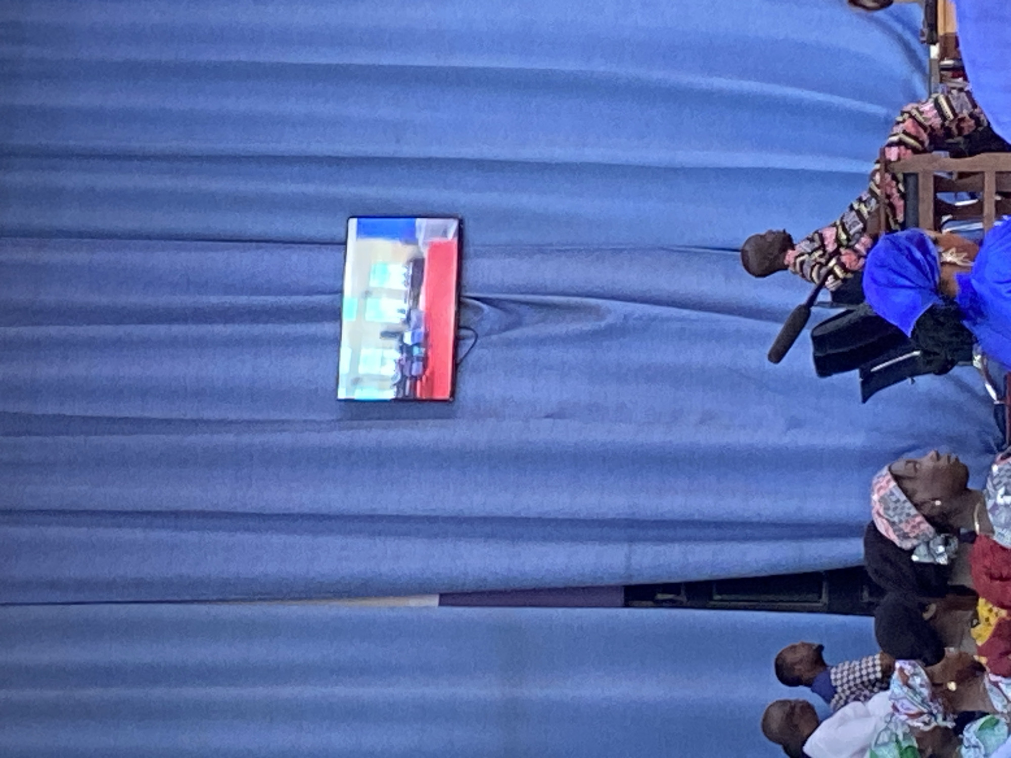 The image displays an indoor scene, specified as being in Mararaba, Nigeria, dominated by an expansive, pleated blue curtain serving as the backdrop. A flat-screen monitor is mounted on the blue curtain, positioned slightly left of the image's vertical center. The monitor displays an interior room, appearing to be a conference or meeting space, furnished with tables covered in red cloths and multiple chairs.
In the mid-ground, on the right side of the frame, a dark-skinned individual with a closely cropped head is seated in profile on a light-colored wooden chair, facing right. This individual is wearing a long-sleeved garment featuring a multi-colored, abstract geometric pattern. A black boom microphone extends from the left, with its head positioned in front of and slightly above the seated person. Behind this individual, two black office chairs are partially visible.
On the left side of the mid-ground, two dark-skinned men are partially visible. The leftmost man, seen from behind and to the side, wears a white long-sleeved shirt. The second man, to his right, is seen in profile and wears a checkered shirt.
The foreground is occupied by a group of individuals, predominantly dark-skinned women, observed from the back or in profile. Many are wearing various head coverings (scarves or turbans) in diverse patterns and colors, including blue, green, and multi-colored designs. Their collective posture suggests they are seated as an audience or attendees at an organized event.