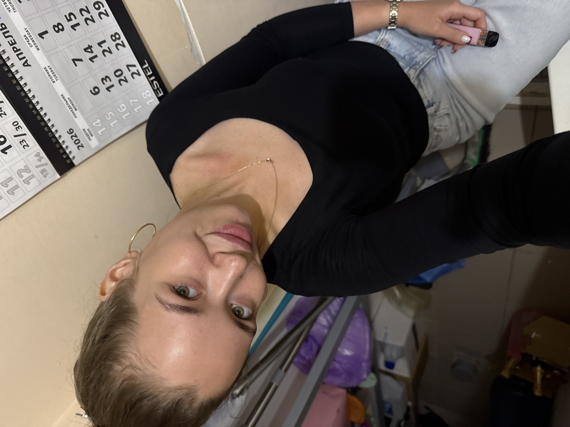 A young woman with blonde hair pulled back and green eyes is taking a selfie indoors. She is wearing a black long-sleeved shirt and light blue jeans. She has a gold necklace and a large gold hoop earring in her left ear. A pink vape pen is visible in her right hand. Behind her, a wall calendar for April 2026 is partially visible, indicating the location is likely Chaikovsky, Russia. The background also shows shelves with various items, suggesting a domestic or storage area.
