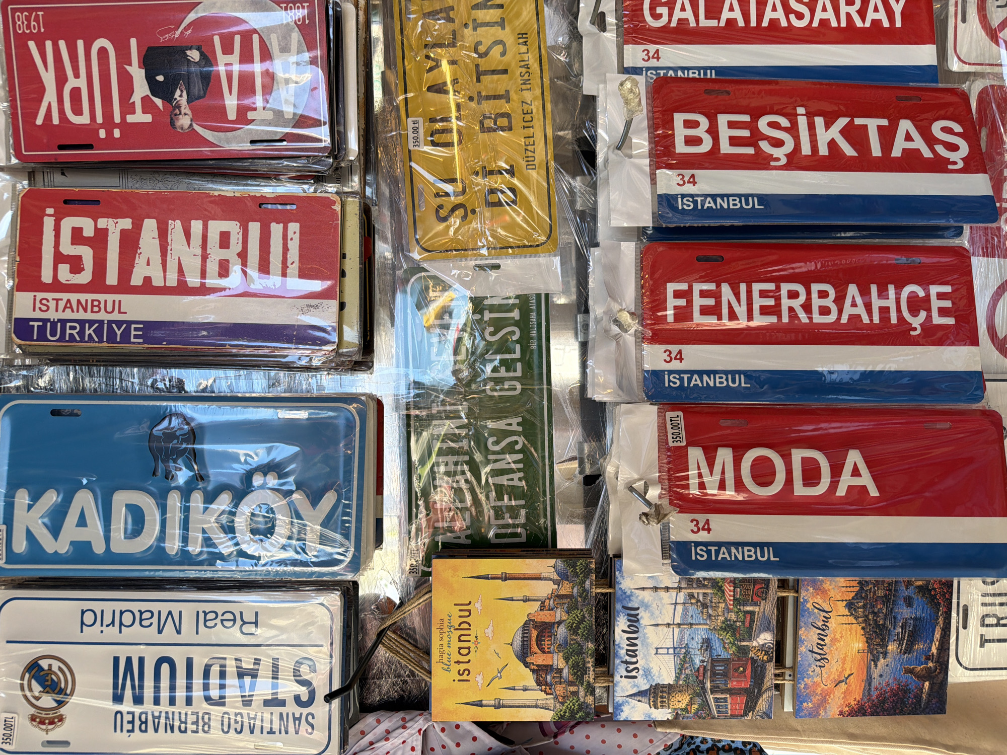 The image displays a close-up, eye-level view of a market stall in Istanbul, Turkey, specifically in the Kadıköy district, filled with souvenir license plates and small decorative items. The dominant objects are numerous colorful license plates, many featuring text and imagery related to Istanbul and Turkish culture. Some plates have "ISTANBUL" prominently displayed, along with neighborhood names like "KADIKÖY," "MODA," "FENERBAHÇE," and "BEŞİKTAŞ." There are also plates referencing famous landmarks like the Hagia Sophia and Bosphorus Bridge. The plates are predominantly red, white, and blue, or yellow. Some are also reproductions of football team logos. In addition to the license plates, there are small square decorative items depicting Istanbul skylines with mosques and bridges. All items appear to be new and individually packaged in plastic. The background is slightly blurred but suggests a busy market environment with other stalls or displays. The lighting is bright, indicating an outdoor or well-lit indoor setting. No people are clearly visible in the frame.
