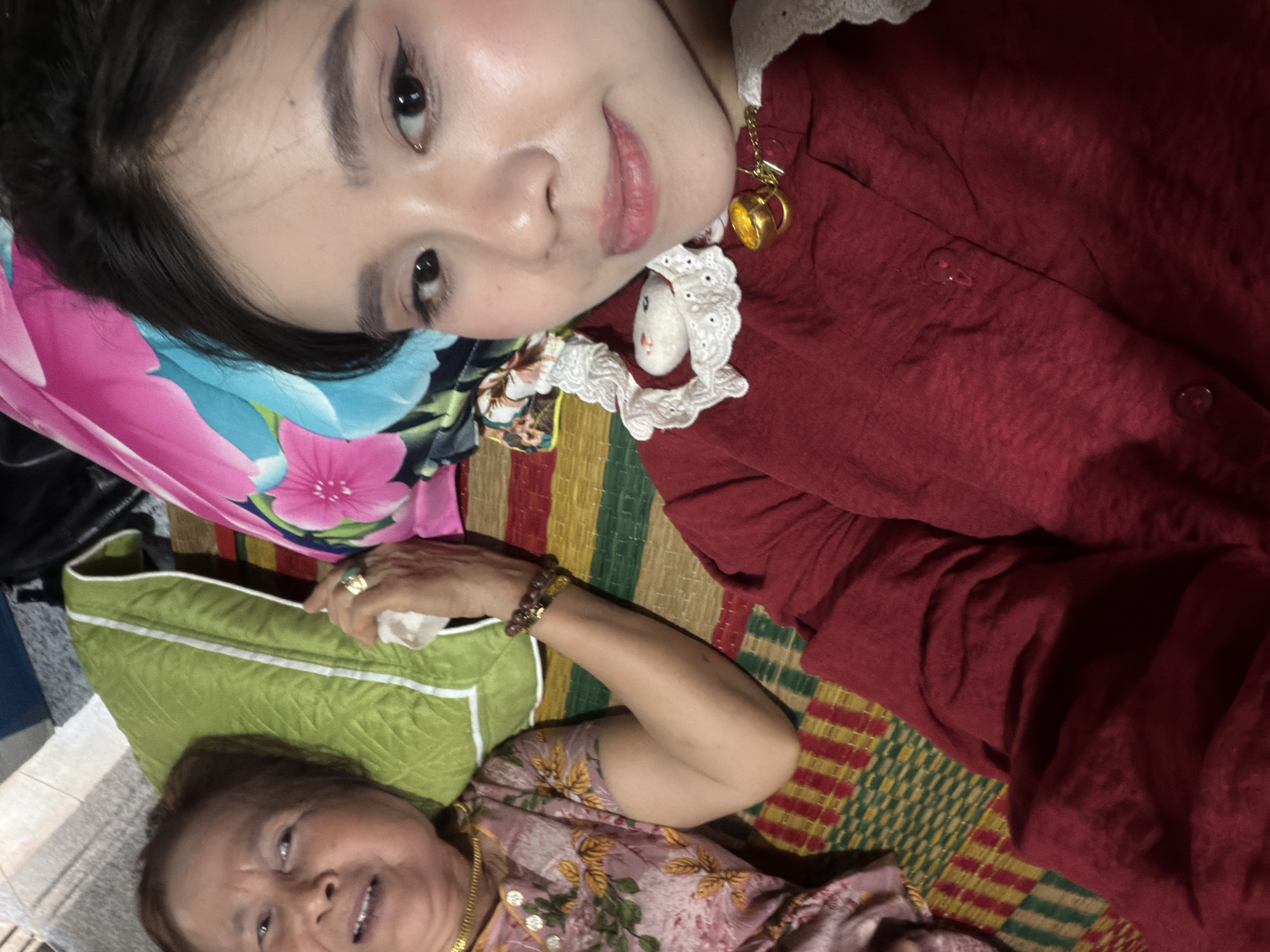Two women are lying on a woven mat in an indoor setting. The younger woman is at the top of the frame, wearing a red shirt with a white lace collar and a necklace with a gold pendant. She has dark hair and is looking at the camera with a slight smile. A floral-patterned cloth, mostly pink and blue, is near her head.
The older woman is at the bottom of the frame, lying on a green pillow with white trim. She wears a pink floral-patterned shirt and a gold necklace. She is also smiling and looking towards the camera. Her arm is visible, with a bracelet and a ring on her hand holding a tissue.
The woven mat underneath is multicolored with a checkered pattern. The background is indistinct. The photograph was presumably taken at the location of Bình An, Vietnam.
