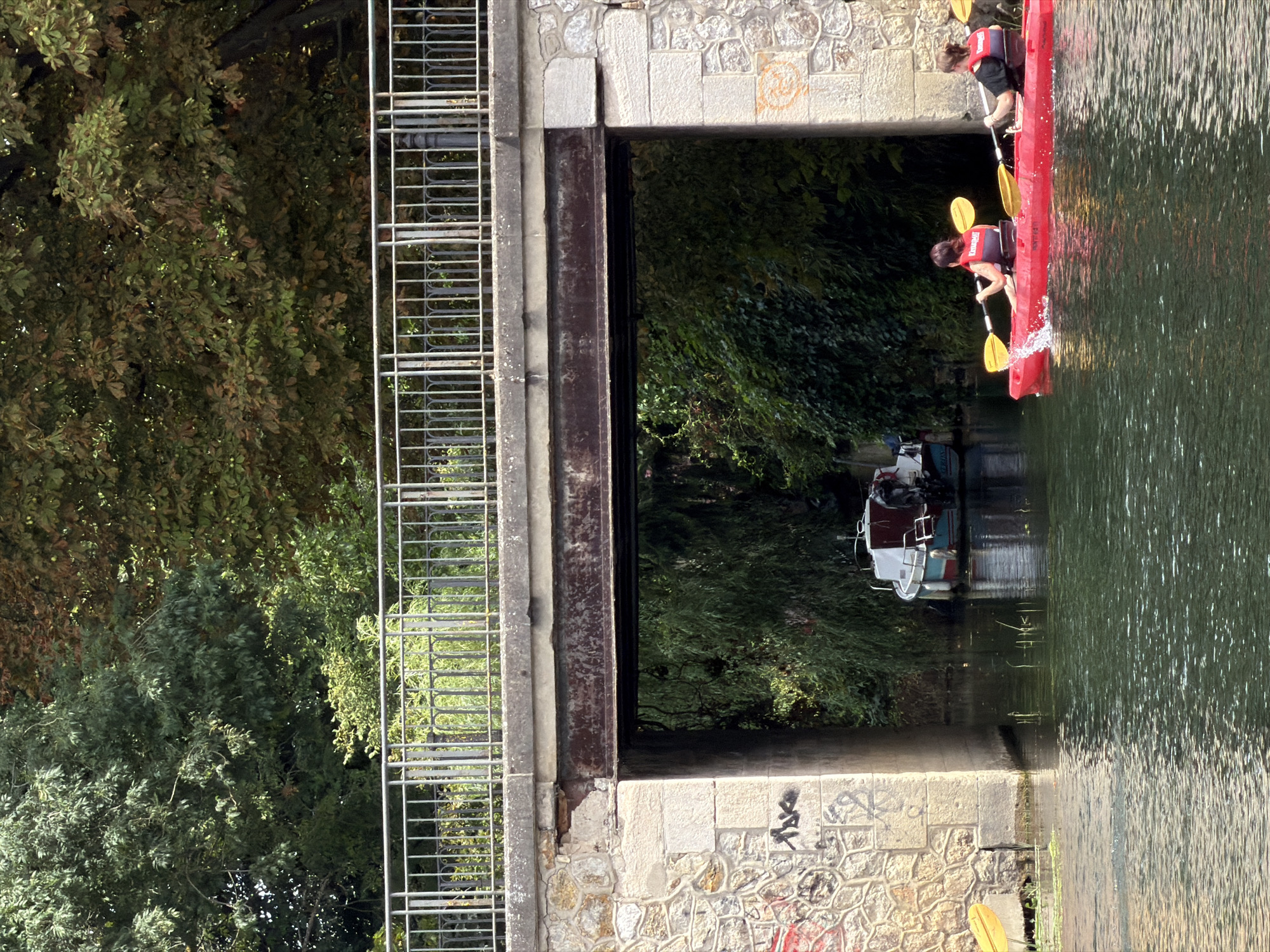 Two individuals are kayaking in a narrow waterway beneath a stone bridge in Joinville-le-Pont, France. The kayaks are red, and both individuals are wearing orange life vests. The person in the front kayak is paddling on the right side, causing a splash of water. The person in the back kayak is paddling on the left side. The bridge has a metal railing and stone supports. Graffiti is visible on the left stone support. A small boat is moored in the waterway to the left of the bridge. Dense green foliage lines the banks of the waterway.