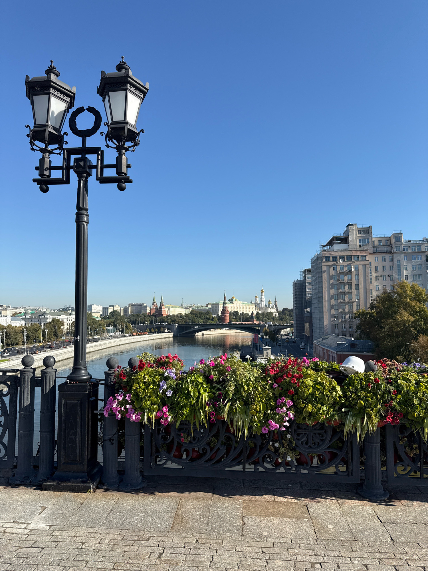This image depicts a view from a bridge in Moscow, Russia, overlooking the Moskva River. In the foreground, a black lamppost with two ornate lamps stands to the left, and a decorative railing adorned with vibrant flower boxes filled with red, pink, and purple blooms lines the edge of the bridge. Beyond the railing, the Moskva River flows, with a bridge crossing it in the mid-ground. The background features the cityscape of Moscow, including several prominent buildings with golden domes, characteristic of Russian Orthodox churches, and a large, modern apartment building. The sky is clear and blue. The overall scene suggests a sunny day in an urban environment.