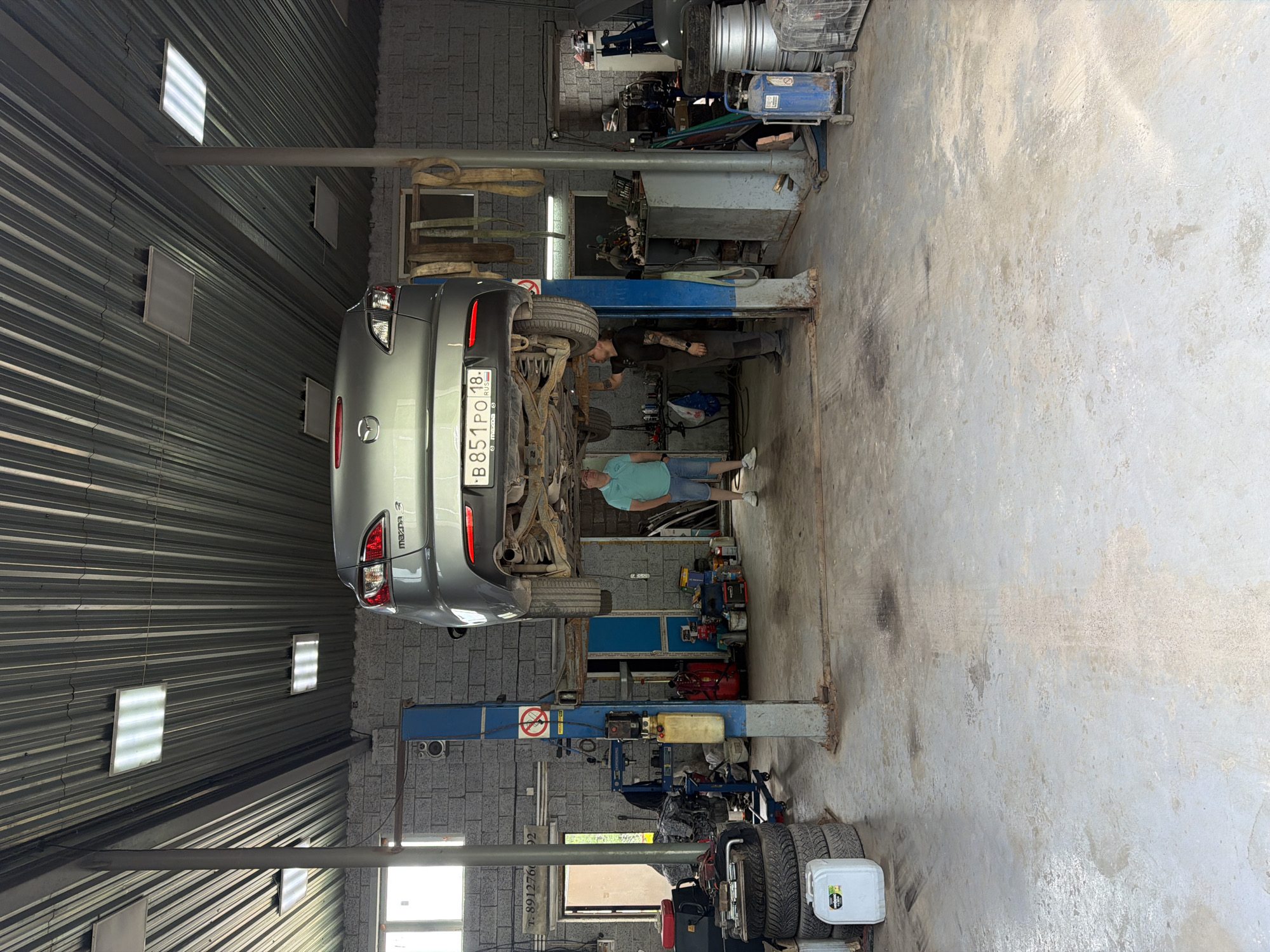 In a car repair shop in Yagul, Russia, a gray Mazda 3 is suspended on a car lift. The vehicle's undercarriage is exposed, indicating ongoing maintenance or repair work. Two men are present in the shop. One man, wearing a light blue polo shirt and denim shorts, stands on the right side of the image, looking towards the car. The other man, dressed in a black t-shirt and dark pants, is positioned beneath the vehicle, appearing to work on it. The shop environment is cluttered with car parts, tools, and equipment, typical of an auto repair facility. Tires are stacked in the foreground, and various containers and machinery are visible throughout the space. The concrete floor shows signs of wear and oil stains. The lighting in the shop appears to be a mix of fluorescent overhead lights and natural light filtering in from an open doorway or window.