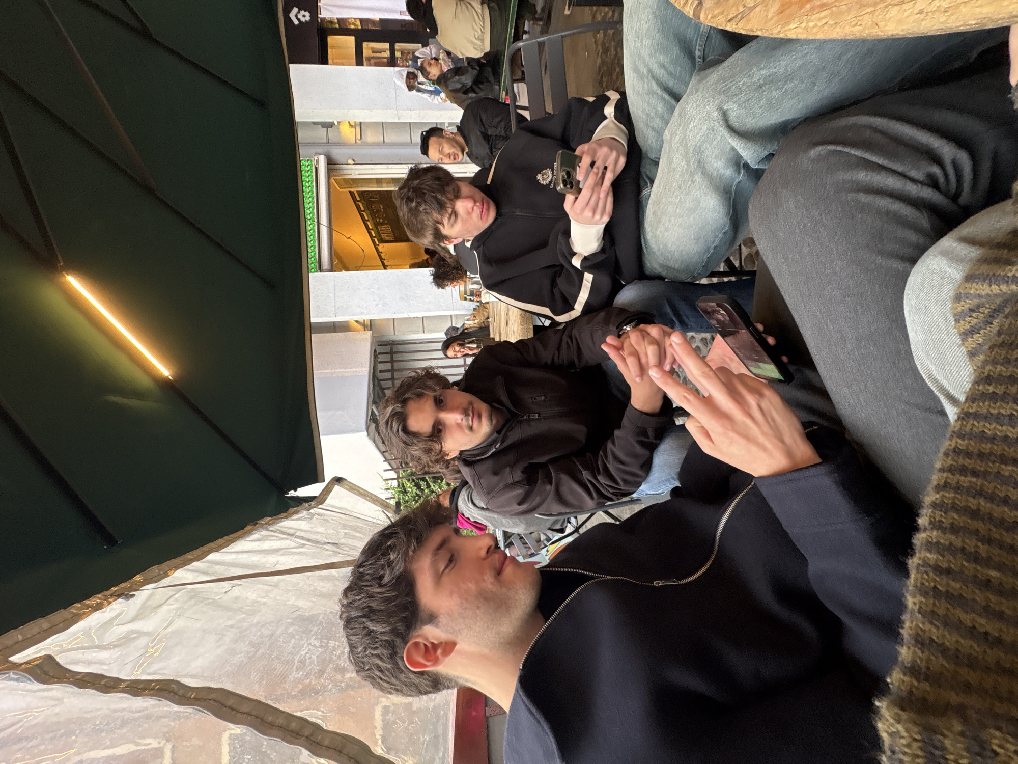 The image shows a group of young adults seated outdoors, likely at a cafe or restaurant in Milan, Italy. The location is suggested by the architectural style of the surrounding buildings and storefronts visible in the background. The people are casually dressed, with several holding and looking at their smartphones. The scene appears to be a candid shot of a social gathering under an awning. The lighting is a mix of natural daylight and artificial lighting from the storefronts.