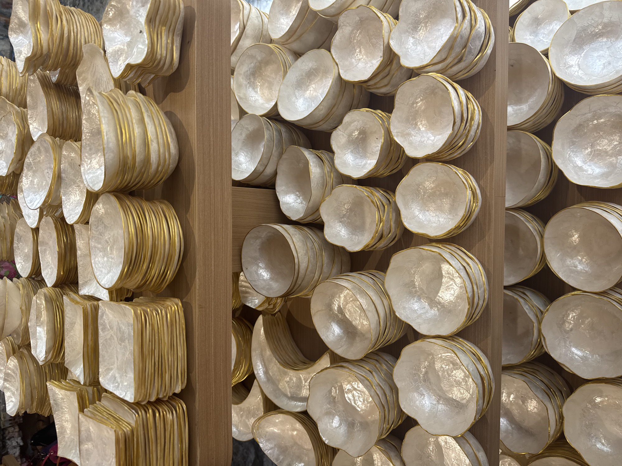 The image depicts a wall decoration made of stacked, pearlescent bowls or plates with gold-colored rims. The decorations are arranged in vertical columns, separated by wooden panels. The objects vary slightly in shape, with some being round and others more square. The lighting is bright and even, highlighting the texture and sheen of the pearlescent material. The location is Kampung Toyapakeh, Indonesia.