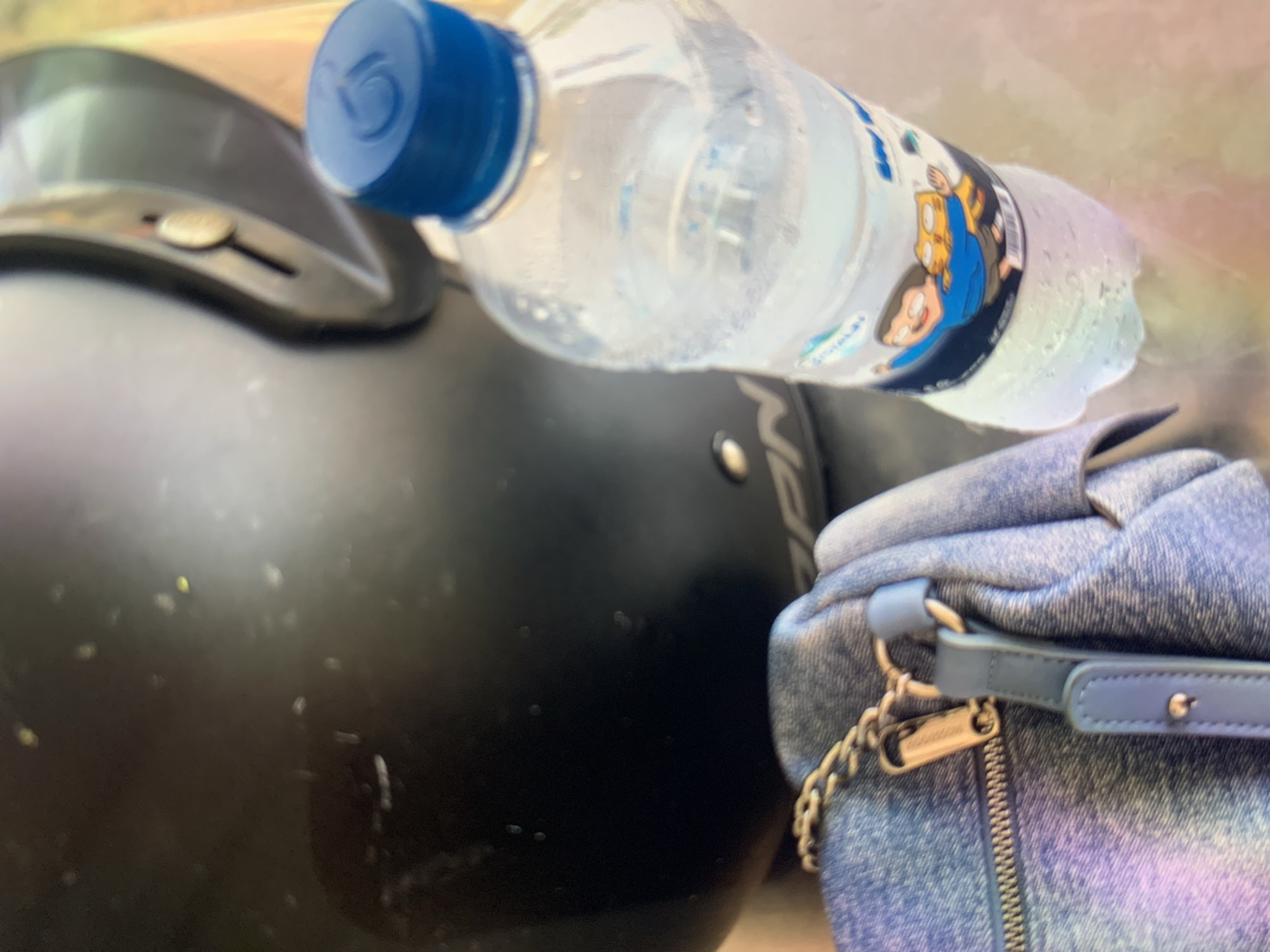A black helmet is positioned on the left side of the image, with a portion of a blue-capped water bottle to its right. The bottle appears to be sweating, indicating it is cold. A denim bag is visible in the lower left corner. The scene is outdoors, with a blurred background suggesting a natural or park-like setting. There are no people or animals present in the frame. The overall lighting suggests it is daytime. The image does not provide any specific clues to pinpoint its exact location within Ciamis, Indonesia.