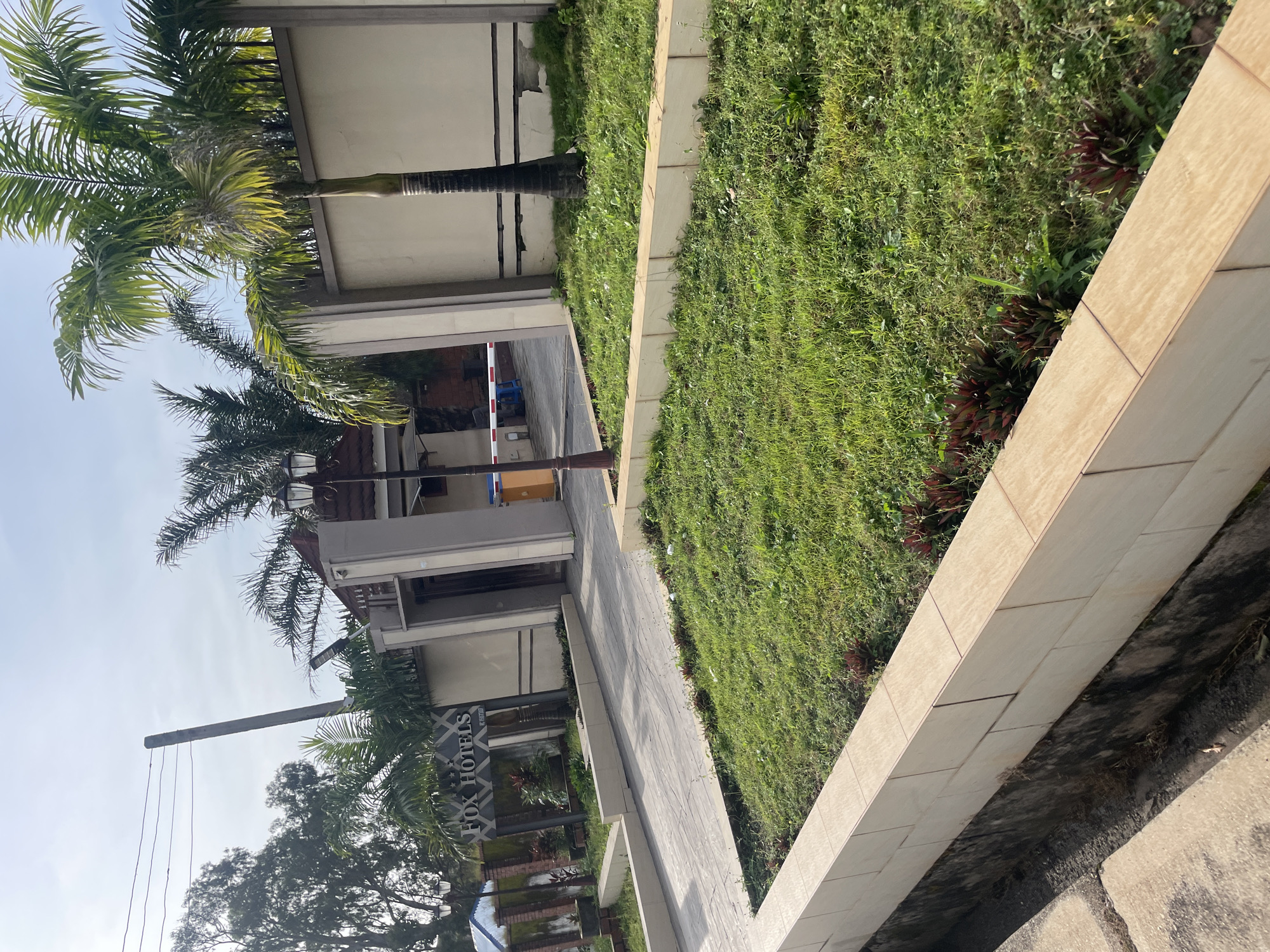 The image shows the entrance to "FOX HOTELS" in Jos, Nigeria. The hotel entrance features a paved walkway leading to a gated entrance with a white and red barrier. On the left side of the walkway, a sign displays "FOX HOTELS" with decorative elements. The entrance is flanked by walls and lush green grass areas, with palm trees and other vegetation visible. A lamppost stands near the entrance. The sky is partly cloudy.