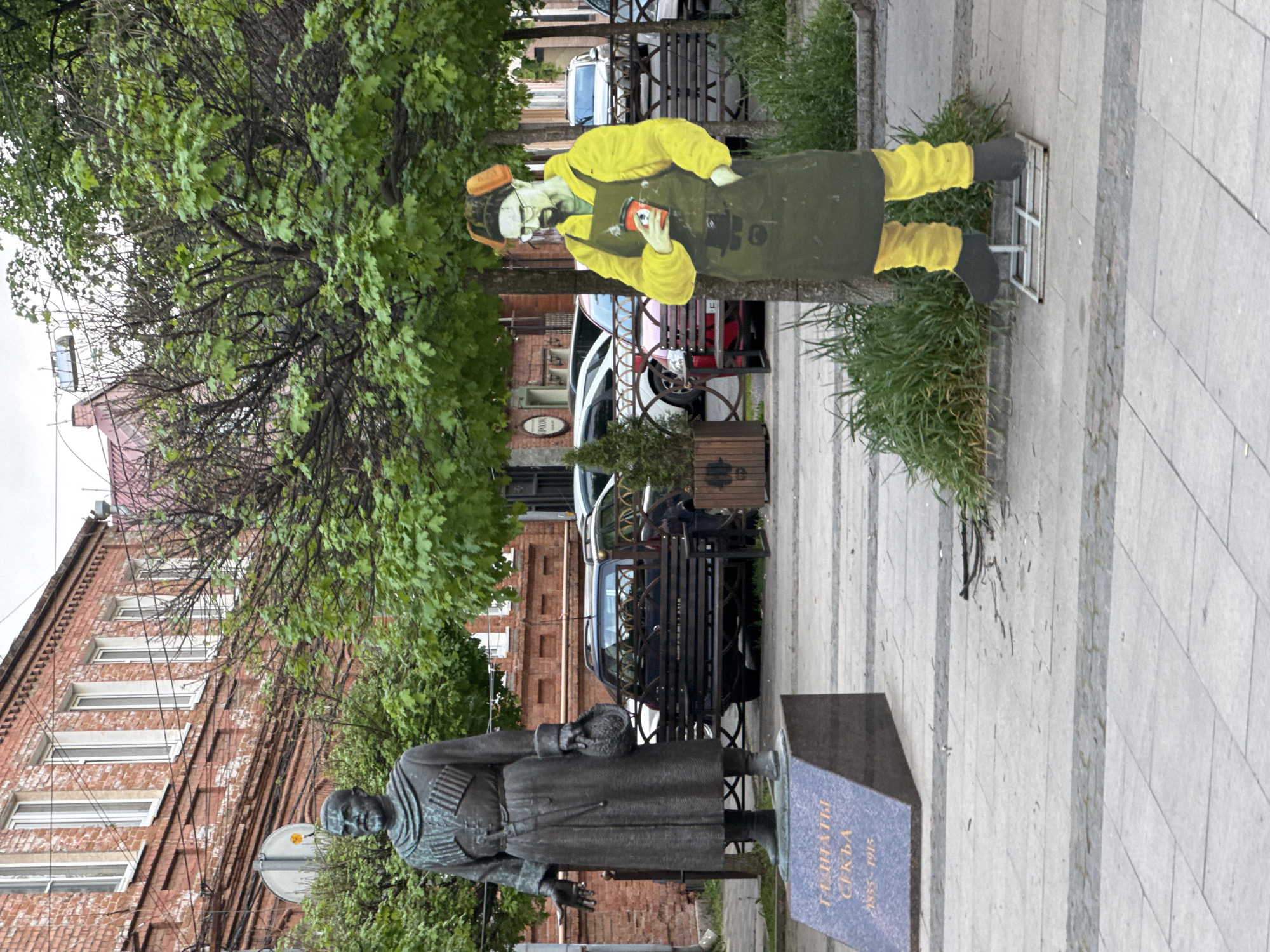 In Akhieli, Georgia, a bronze statue of a man in a long coat stands near a tree and a painted figure of a man in a yellow outfit and dark apron holding an object. A brick building and parked cars are in the background. The area is paved with light gray stones.
