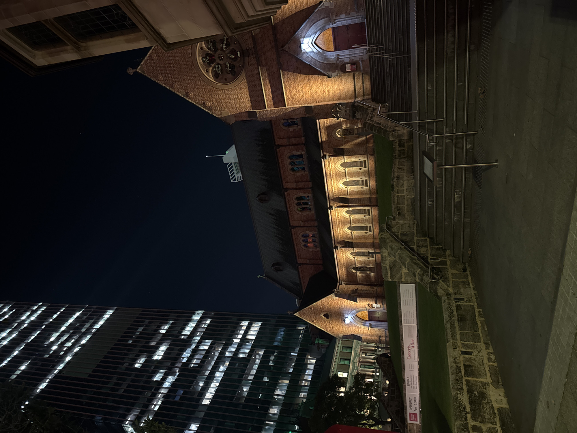 The image captures a nighttime urban scene in Perth, Australia, showcasing a striking contrast between historical and modern architecture. On the right, a grand, old brick cathedral, likely St George's Cathedral, dominates the frame. Its intricate brickwork, arched windows, and a prominent circular rose window are warmly illuminated by artificial lights, casting an inviting glow. Steps lead up to its main entrance, and a dark, multi-faceted roof with a spire is visible against the night sky. A robust stone wall borders a small grassy area in front of the cathedral.
To the left, a tall, modern glass skyscraper stands, its numerous windows emitting bright, cool white light, creating a grid-like pattern that reflects the city's contemporary pulse. The dark, clear night sky provides a dramatic backdrop for both structures, emphasizing their distinct lighting and architectural styles. The foreground features a paved ground area and the aforementioned grassy patch.
Visible text on a sign next to the cathedral reads "Concerts At One" and includes details like "SUNDAY" and "OPEN DAILY" with times "7pm - 5.30pm", indicating events and opening hours. Partial text for "St George's Cathedral" is also visible, confirming the building's identity. The scene is devoid of people, contributing to a serene and majestic atmosphere that highlights the architectural beauty and history of the location after dark.