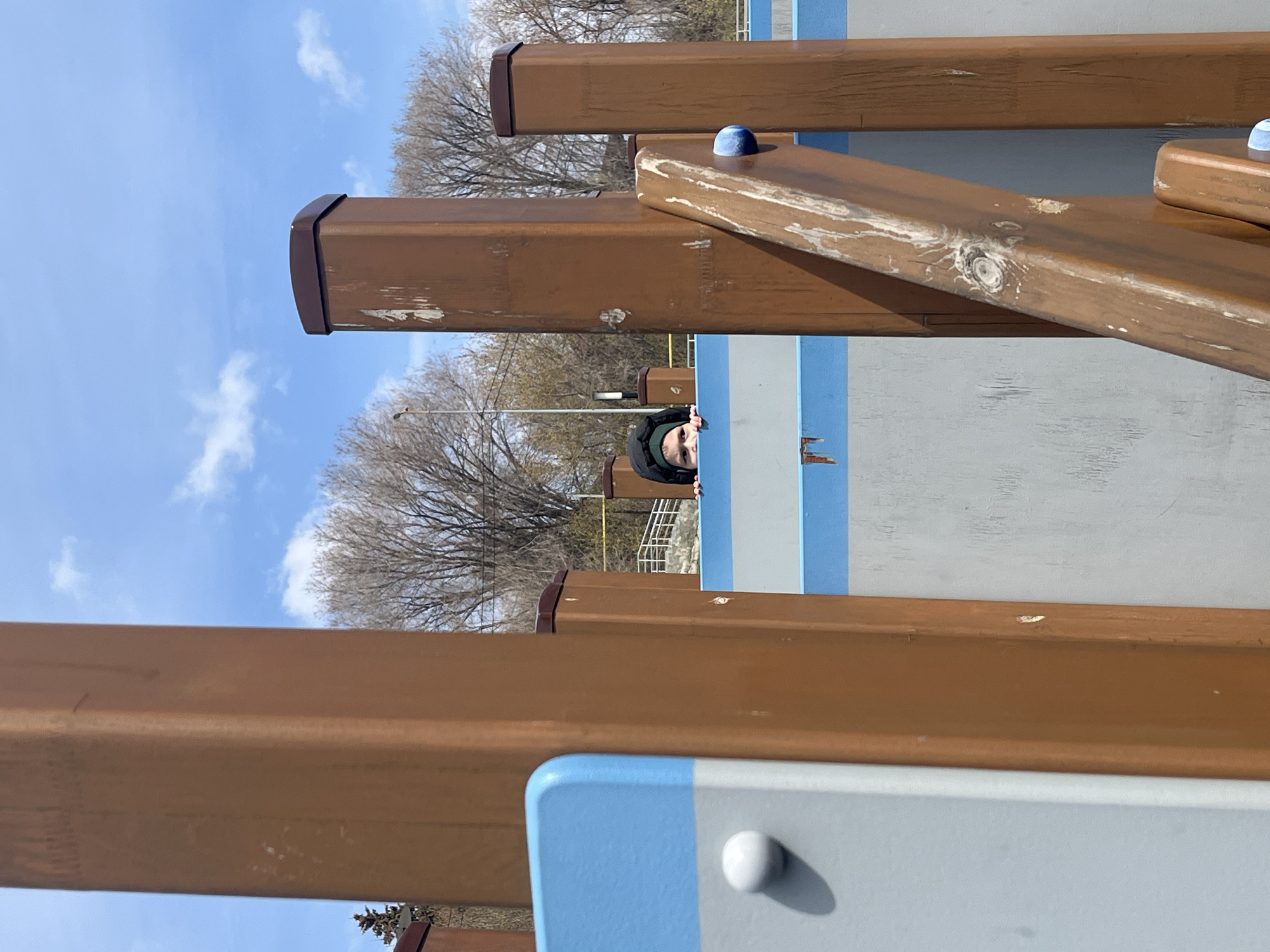 The image captures an outdoor scene, likely a playground or park, featuring a child peeking from behind a structure. A young person, wearing a dark hooded jacket, is partially visible, with their eyes and forehead appearing over a light blue and grey panel on the right side of the frame.
The foreground and midground are dominated by several large, rectangular beams, which appear to be made of wood or a wood-like material. They are a warm brown color, with noticeable wear, scratches, and lighter streaks, showing natural wood grain patterns in some areas. These beams are arranged vertically and horizontally, forming part of a play structure. Small, spherical elements, one blue and one white, are integrated into the structure, suggesting interactive components.
In the background, a bright blue sky with scattered white, wispy clouds is prominent. Several deciduous trees with bare branches are visible against the sky, indicating late winter or early spring. The lighting suggests a clear day. No visible text is present in the image. The overall atmosphere is playful and bright.