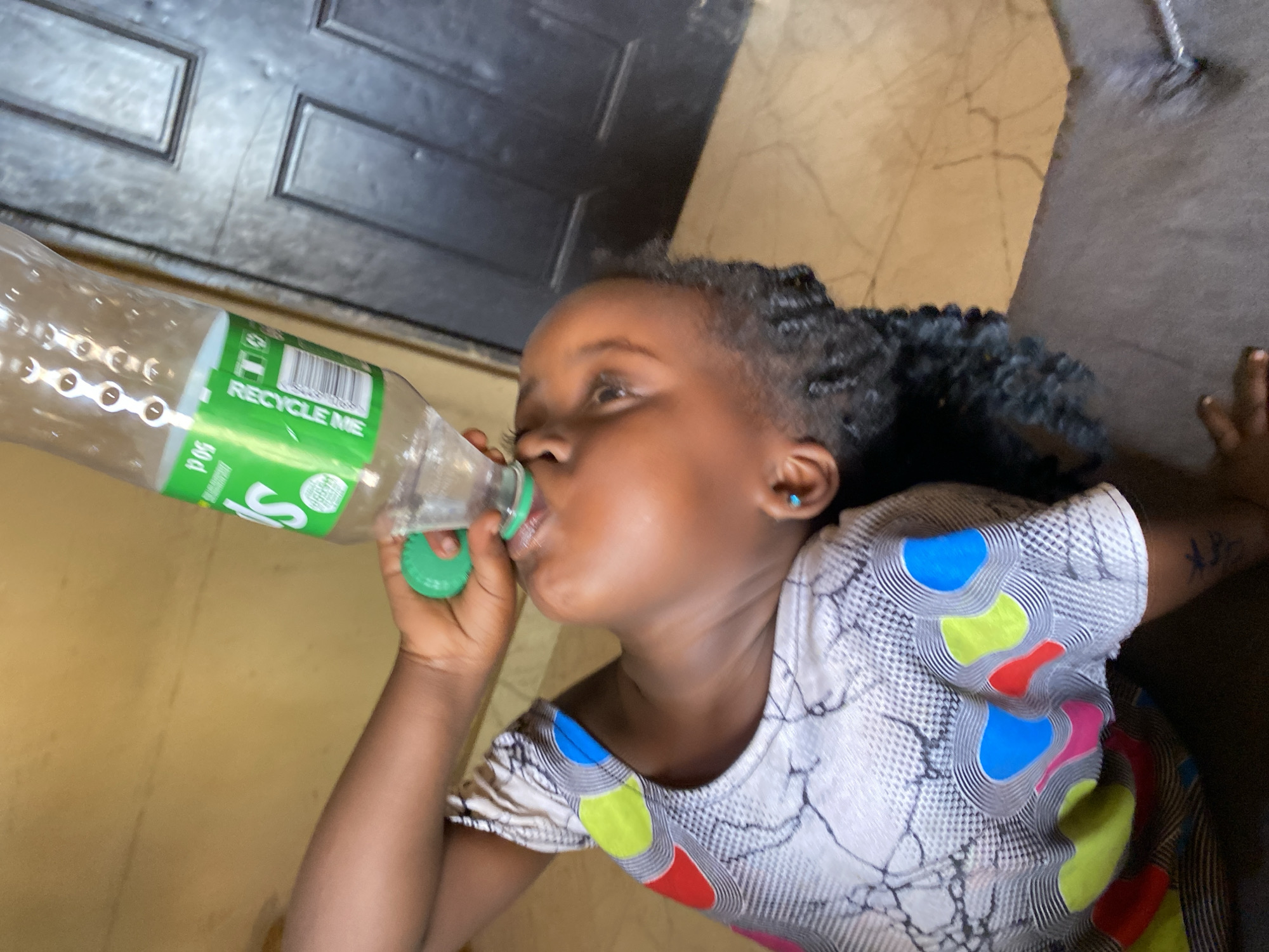 A close-up, slightly angled shot captures a young child, likely a girl, drinking from a clear plastic bottle with a green cap and label. The child has dark skin and braided hair, wearing a small blue stud earring in her right ear. Her attention is focused on the bottle as she sips. She is dressed in a light-colored shirt featuring a intricate pattern of black lines and distinct, vibrant abstract shapes in blue, lime green, red, and pink. Her arm is visible, holding the bottle.
In the background, a dark-colored door with two recessed panels is partially visible to the upper left. The floor appears to be a light-brown or beige tiled surface. The lighting suggests an indoor setting during the daytime in Kuru Dungus, Nigeria.
Visible text on the bottle's green label prominently includes "RECYCLE ME," along with "50cl" indicating volume. A barcode and other small, illegible symbols are also present on the label.