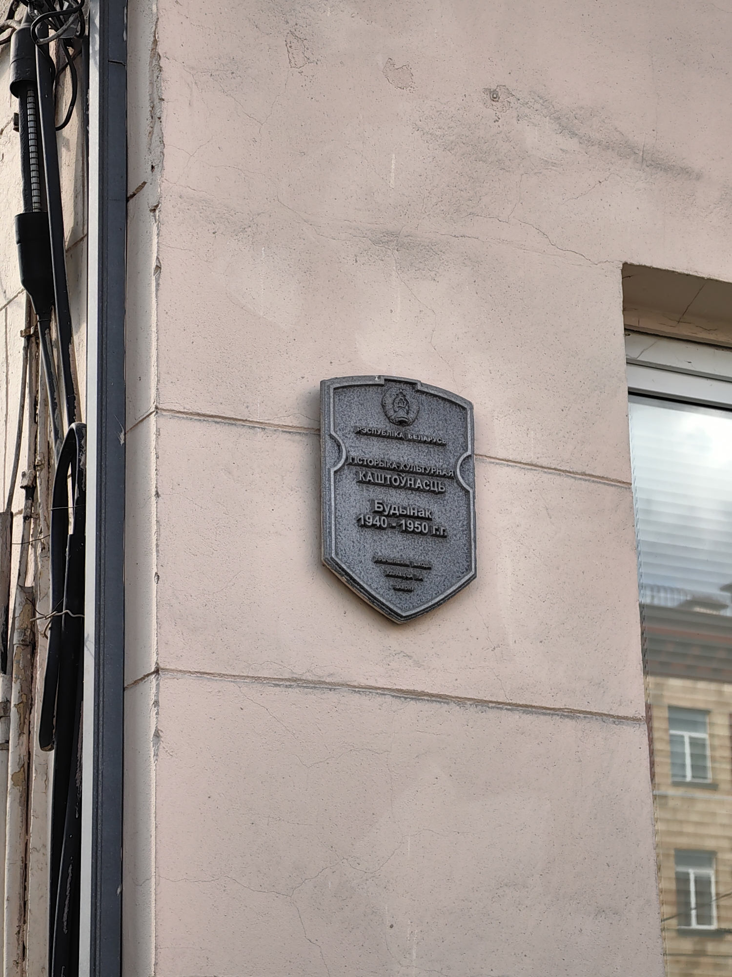 This media file shows a close-up view of a building with a commemorative plaque on its exterior wall. The plaque is metallic and shield-shaped, mounted on a light-pink, textured wall. The wall shows signs of age, with some cracks and discoloration. To the left of the plaque, a section of a building's facade is visible, featuring wiring and conduits running vertically. A window with horizontal blinds is also visible on the right side of the image, reflecting another building with windows.
The plaque itself contains text in Cyrillic script. At the top, it reads "РЭСПУБЛІКА БЕЛАРУСЬ" (Republic of Belarus), beneath which is an emblem. Below the emblem, the text states "ГІСТОРЫКА-КУЛЬТУРНАЯ КАШТОУНАСЦЬ" (Historical and Cultural Value). The central inscription reads "Будынак 1940-1950 гг." (Building 1940-1950). The bottom portion of the plaque has smaller text, likely identifying the original purpose of the building or its significance, reading "ПРЭВЫШЭЙШЫ ШКОЛА" (Higher School) and "КРАСНАЯ ПЛ." (Krasnaya Pl.).
The scene appears to be outdoors, likely during daylight hours, given the lighting. The overall impression is one of urban architecture with a focus on historical preservation. The plaque indicates that the building has historical and cultural significance and served as a higher school between 1940 and 1950. The location context provided is Minsk, Belarus.
