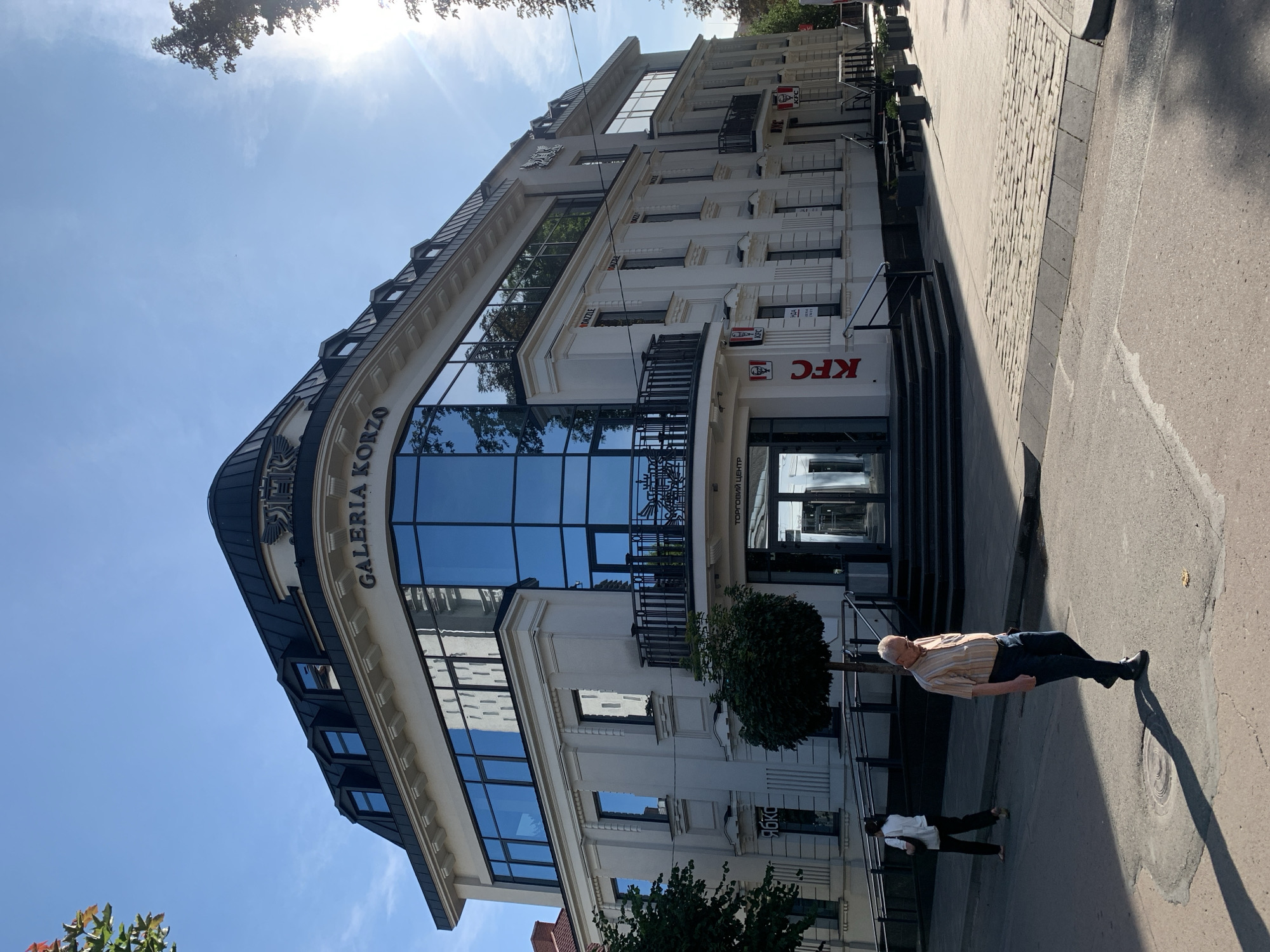 This image captures a sunny daytime exterior view of the "Galeria Korzo" shopping center, located in the city of Kolomyia, Ukraine. The building is a multi-story structure with a light-colored facade and a dark, curved roofline featuring dormer windows. Large, reflective blue windows dominate the upper floors, displaying reflections of trees and the sky. A prominent sign on the facade reads "GALERIA KORZO".
At street level, the building houses multiple establishments, including a KFC restaurant, identifiable by its red and white logo and signage. The entrance to the shopping center is marked by a set of stairs. In the foreground, two individuals are walking on the paved street. An elderly man in a striped shirt and dark pants is walking towards the right, casting a long shadow. Further back and to the left, a woman in a white shirt and dark pants is walking away from the camera. The street is paved with asphalt and cobblestones in sections. The environment appears to be a city street with trees lining the sidewalks and buildings in the background. The sky is clear and blue with a bright sun casting strong shadows.