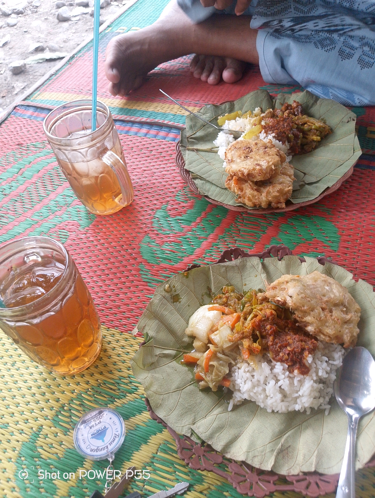 Two plates of food are presented on a woven mat. Each plate consists of white rice, a fried item, and a side dish with vegetables and chili sauce, all served on large green leaves. Two glasses of iced tea are also on the mat. In the background, a person's bare feet are visible, and a blue straw extends from one of the glasses. The scene appears to be outdoors on a mat, possibly in Slawi, Indonesia. A keychain with text, including "Temu kangen" and "PTM," is also visible near one of the glasses. The image has a timestamp indicating it was "Shot on POWER P55."