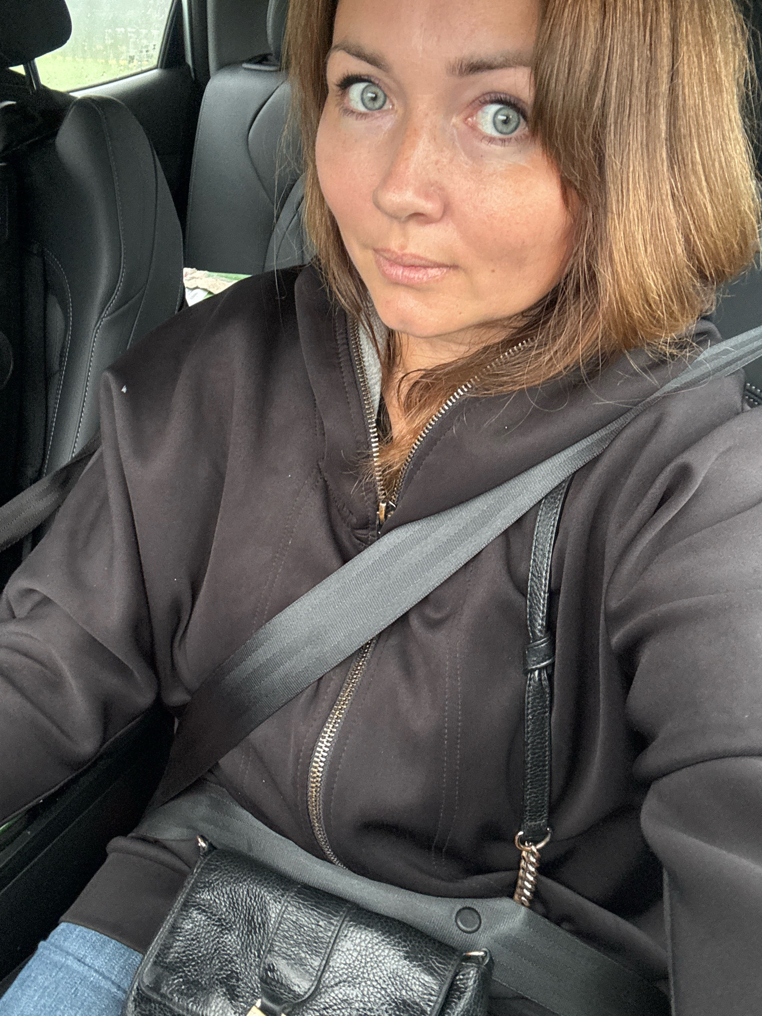 A woman is seated in the driver's seat of a car, as indicated by the presence of a seatbelt. She is wearing a black zip-up hoodie and jeans. A black leather purse with a chain strap is visible, worn across her body. The background shows the interior of a car with black seats. The location is identified as Rappolovo, Russia.