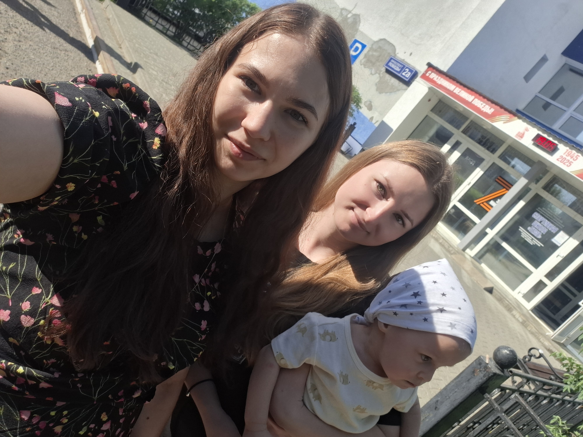 Three people are visible in the image, apparently taking a selfie outdoors. A young woman with long brown hair and wearing a black floral dress is holding the camera. Next to her is a second young woman with blonde hair. A baby is being held by one of the women. The baby is wearing a white shirt with animal prints and a white headscarf with gray stars. Behind them is a building with the address "проспект победы 2а" indicating this is in Chaikovsky, Russia. The building is decorated with "с праздником великой победы!" indicating a Victory Day celebration, and there is a symbol that appears to be a 'Z' made out of St. George's ribbon. The display behind the ribbons shows '9:05 1945 2025' which is likely for the 80th anniversary of Victory Day in 2025.