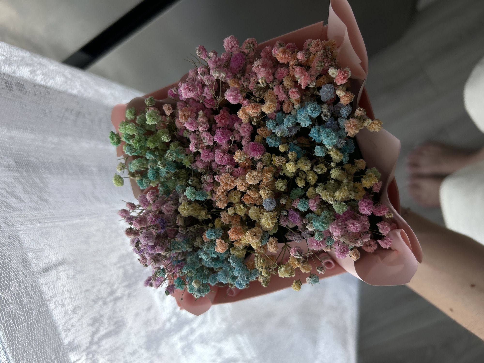 An overhead shot captures a vibrant bouquet of dried baby's breath flowers, dyed in an array of pastel colors including pink, blue, green, yellow, orange, and purple. The small, delicate flower clusters create a rich, textured surface. The bouquet is neatly wrapped in light pink paper, which complements the floral tones. A person's bare foot is partially visible in the lower right, and a forearm or hand can be seen on the right edge, indicating someone is holding or presenting the flowers.
The bouquet is positioned above a white, translucent fabric with a subtle, textured pattern, resembling lace or a sheer curtain, allowing light to filter through. The background is softly blurred, suggesting an indoor setting illuminated by natural daylight, as evidenced by shadows and bright areas. A dark, indistinct element is visible in the upper background, contrasting with the lighter tones of the foreground. The overall scene evokes a cheerful and gentle mood. No text is visible in the image.