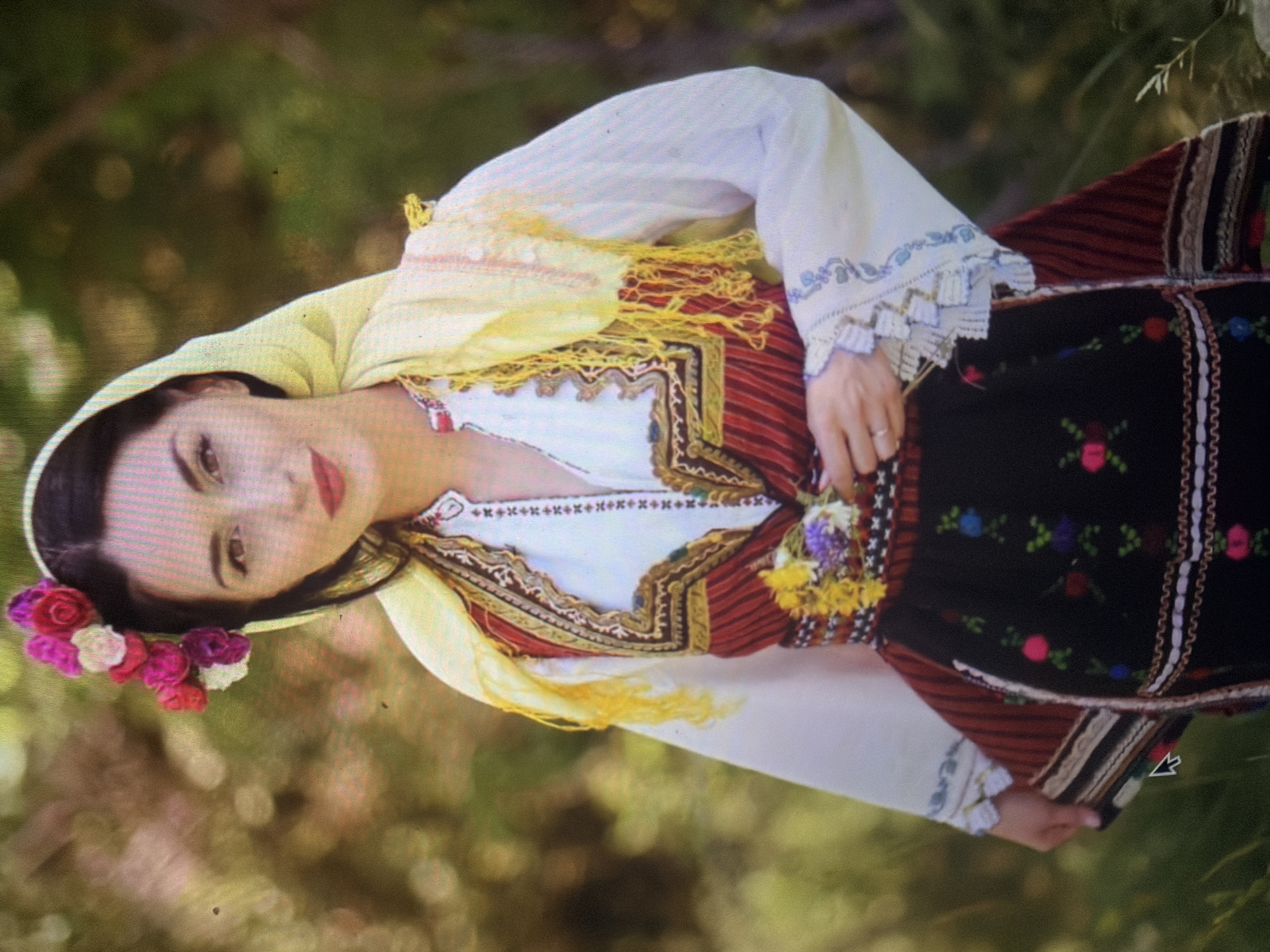 The scene is situated in Shumen, Bulgaria. A single female subject, presenting with dark hair, dark eyes, and red-painted lips, stands facing the viewer with a subtle smile. She is adorned in traditional Bulgarian folk attire. A yellow fabric head covering is draped over her head and shoulders, featuring a cluster of pink, fuchsia, and white artificial flowers attached above her left temple. She wears a white long-sleeved blouse with ruffled, lace-trimmed cuffs embroidered with light blue patterns, and a central vertical embroidered strip on the chest detailed with red and white motifs. Over this, she wears a sleeveless, dark red garment or vest with extensive gold and brown patterned embroidery along its edges and front opening. A multi-colored woven sash, predominantly featuring yellow and brown tones, is cinched around her waist, from which a small cluster of yellow and purple wildflowers protrudes on her left side. A black apron, adorned with multicolored floral and green leaf embroidery and white geometric patterns, covers a vertically striped red and dark fabric skirt. Her left hand, adorned with a ring on the fourth finger, gently holds the fabric of her skirt near her hip. The background consists of a blurred, natural outdoor environment, characterized by green and light brown foliage and soft lighting.
