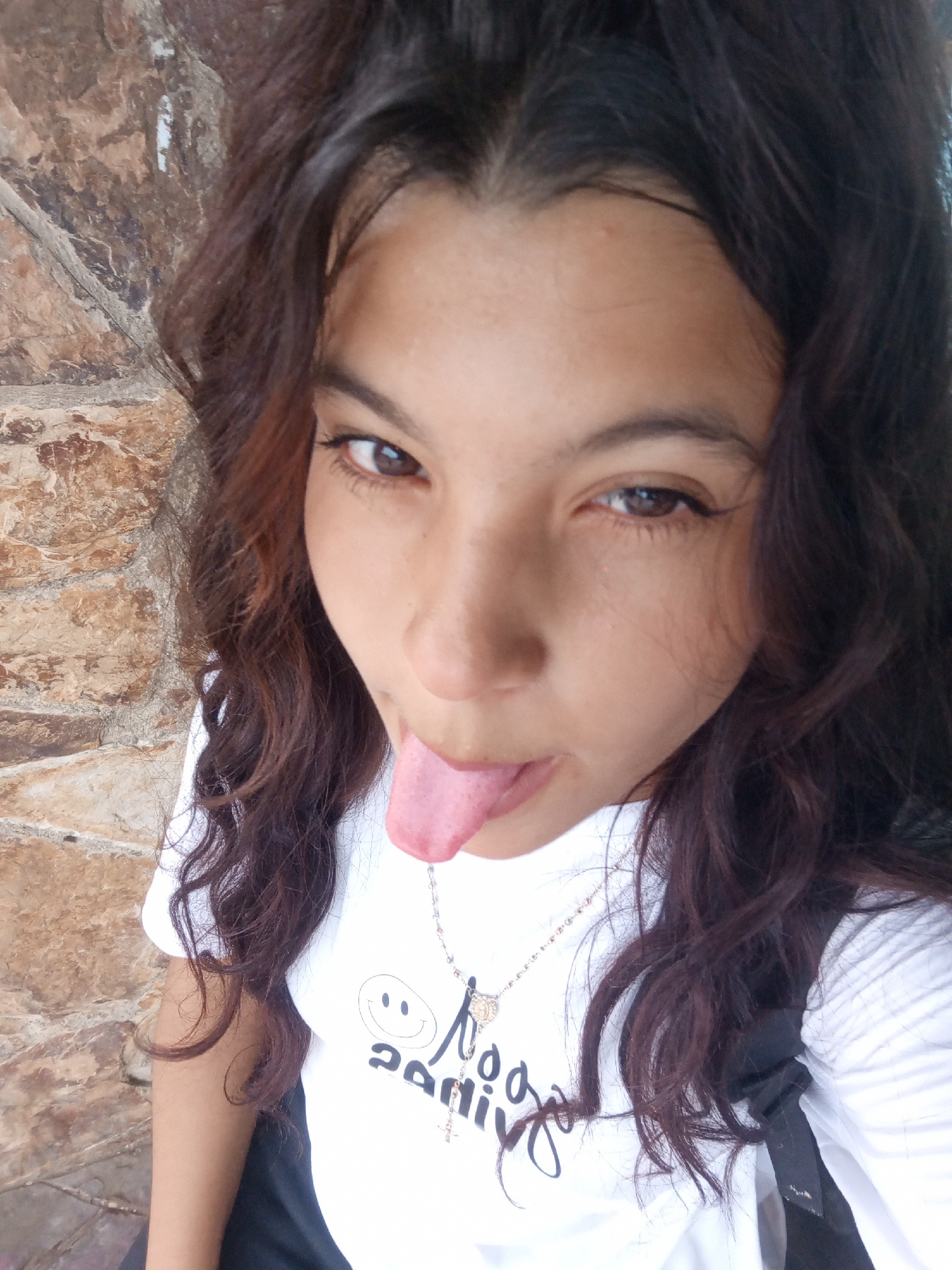 A young woman with dark curly hair is facing the camera, sticking her tongue out. She is wearing a white t-shirt with a smiley face and the word "sad" printed on it. She is also wearing a necklace with a pendant. The background appears to be a wall made of stone or brick. There is no specific information available about this image being taken in the city of Altagracia, Venezuela, or any forensic information.