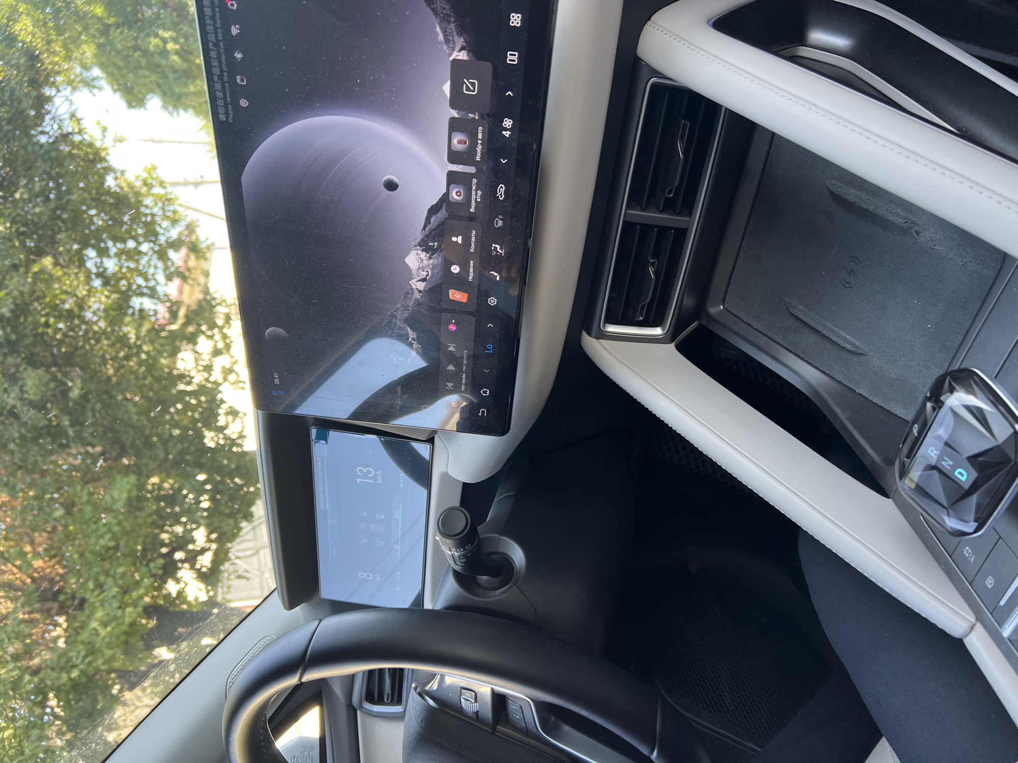 An interior shot of a modern car is captured, focusing on the dashboard and center console. The view is from the passenger seat, looking towards the driver's side. A large, sleek touchscreen dominates the center of the dashboard, displaying a cosmic-themed wallpaper with planets and mountains. Below the touchscreen, a digital instrument cluster is visible, showing speed at 13 km/h and power at 8 kW. The steering wheel, made of black leather, is partially visible on the left. A gear shifter with illuminated letters (P, R, N, D) is prominently placed on the center console. To the right of the gear shifter, a wireless charging pad with a lightning bolt symbol is integrated. The car's interior features white and black upholstery. In the background, through the windshield, trees and sunlight are visible, suggesting an outdoor setting in a city during the day. The overall impression is one of advanced technology and minimalist design.