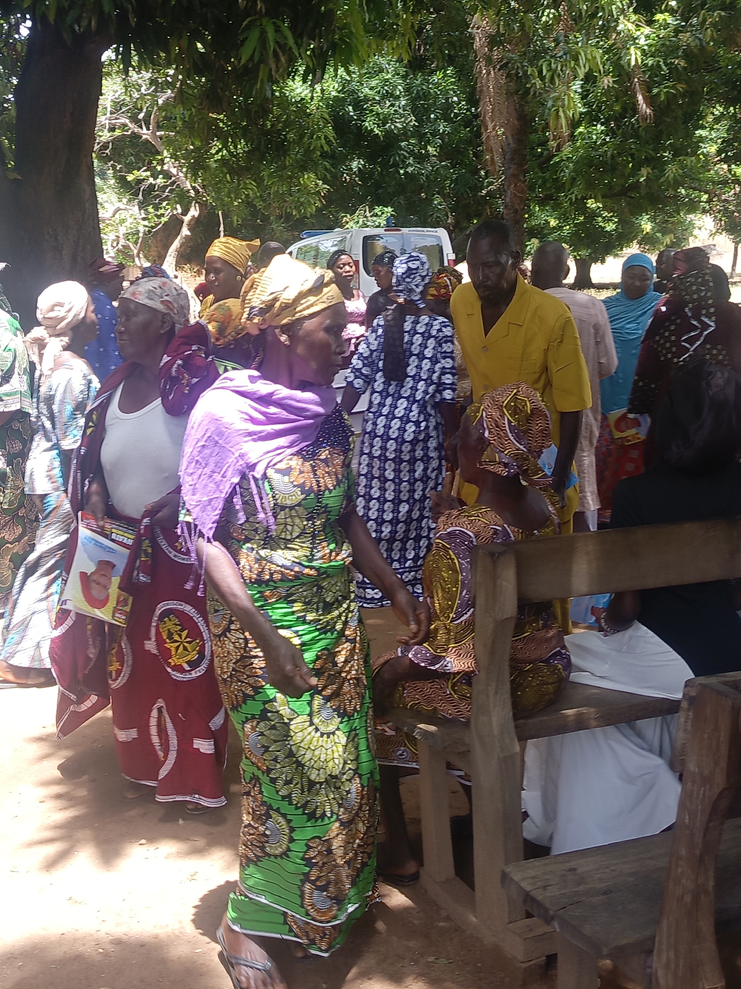 The image shows a group of people gathered outdoors in Ninzo South, Nigeria. They appear to be primarily Black and dressed in traditional or brightly colored clothing, including patterned dresses and headwraps. A man in a yellow shirt stands among them. Some people are standing, while others are sitting on wooden benches. A white van is visible in the background, partially obscured by trees. The environment is shady with trees overhead and the ground appears to be dirt.