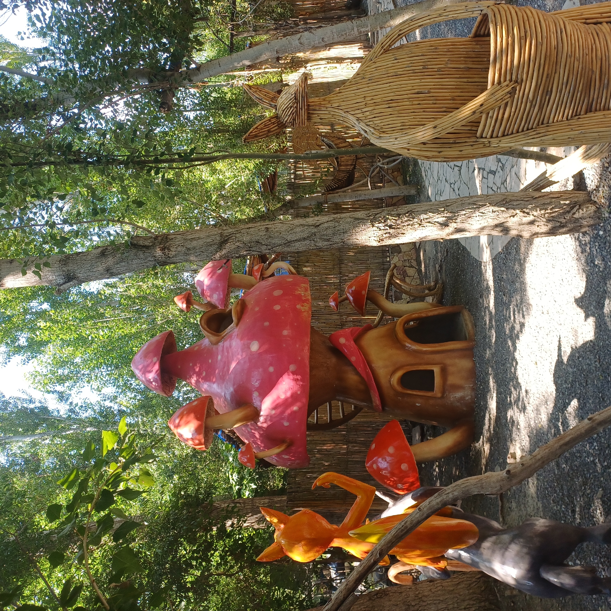 This is a medium shot, eye-level photograph taken outdoors on a sunny day. The scene is a whimsical, garden-like setting with a focus on fantastical, nature-inspired sculptures. In the center of the image is a large, red and pink mushroom house with a whimsical design, featuring multiple mushroom caps and openings resembling windows and doors. To the left of the mushroom house is an orange, cartoonish fox statue with its arms raised. To the far right is a large, woven sculpture of a rabbit or similar animal with long ears, made from light-colored wooden sticks. The ground is covered with small gray gravel, and a stone pathway curves through the scene. The background is filled with lush green trees and foliage, creating a dappled light effect on the ground. The overall impression is one of a playful, artistic outdoor space, possibly a themed garden or a section of a park. There are no people visible in the photograph. The time of day appears to be midday due to the bright sunlight and the shadows cast.