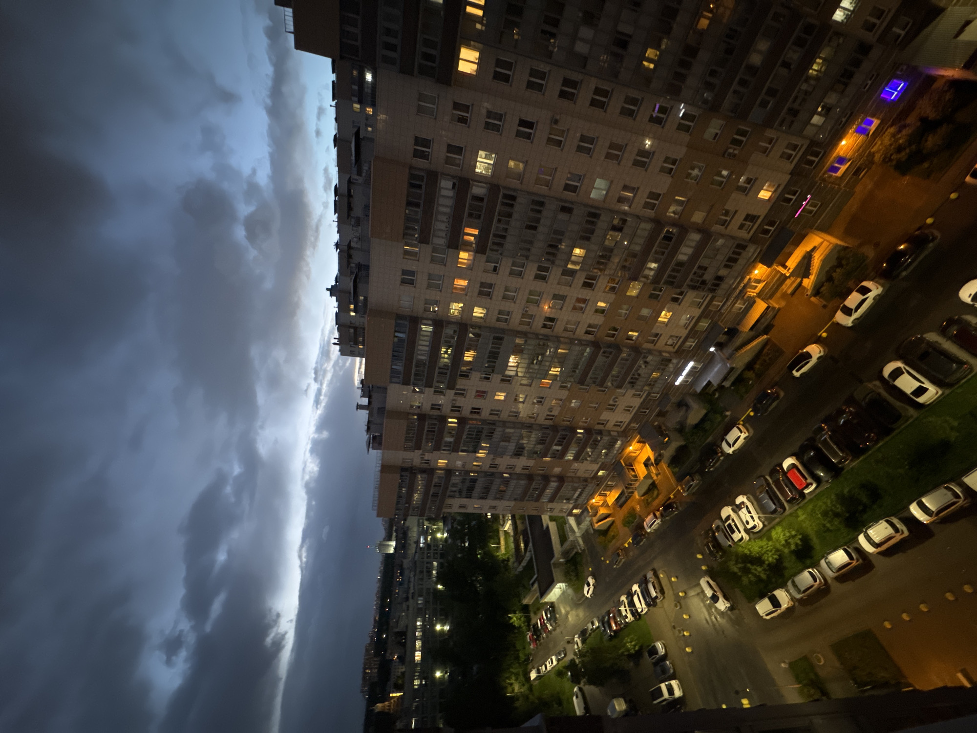 An angled, high-angle view captures a residential area in Sinyavino, Russia, during twilight or evening. On the left, ominous, dark storm clouds dominate a significant portion of the frame. In the center, tall apartment buildings, likely Soviet-era structures, rise vertically, their many windows illuminated with a warm, yellow glow, suggesting inhabitants are home. To the right of the buildings, a street lined with parked cars runs parallel to the apartment blocks. The cars are mostly white, though some black and red vehicles are interspersed. Streetlights cast a warm, yellowish light onto the street and parked cars, adding to the overall contrast between the gloomy sky and the inhabited buildings. In the distance, glimpses of other buildings and green spaces can be seen. The scene lacks any human presence, focusing instead on the interplay of urban architecture, atmospheric conditions, and artificial light.