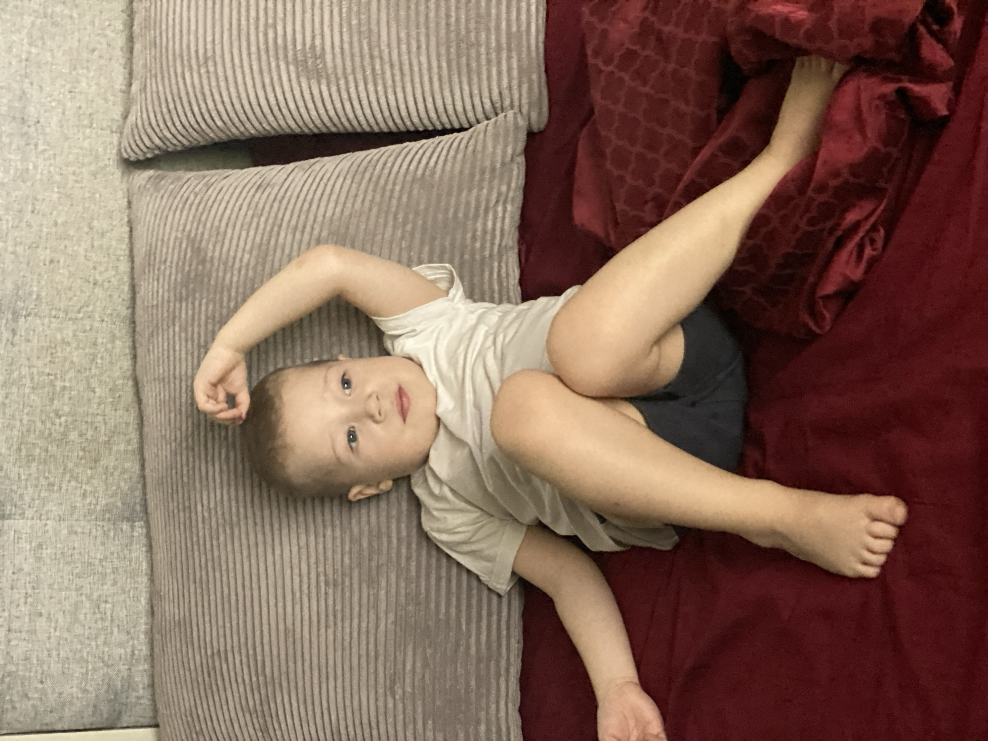 A young child, likely a toddler, lies on a sofa or bed. The sofa or bed appears to be comprised of two distinct sections: a grey, corduroy-textured cushion or section on the left, and a red, patterned, possibly velvet material on the right. The child is positioned diagonally across these sections. The child is fair-skinned, with blonde hair and blue eyes. They are wearing a light-colored t-shirt and dark shorts. The child's right arm is raised with the hand near their head. Their left arm extends down along the red section. The child is looking toward the camera.
There is no indication in the image to suggest this scene is located in Sochi, Russia.