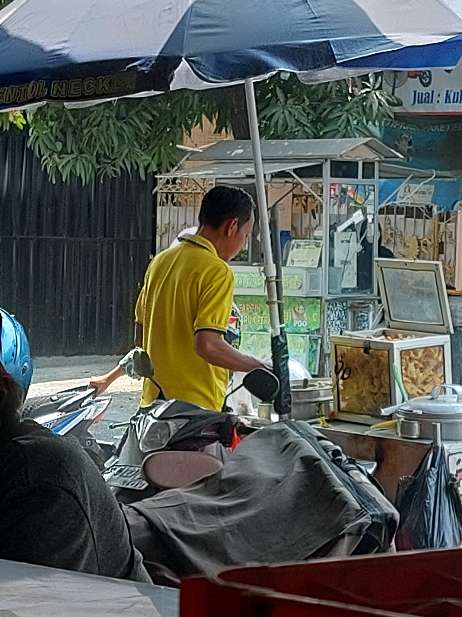 A street scene in Sumuragung, Indonesia, features a man in a yellow polo shirt standing next to a parked motorcycle under a blue and white striped umbrella. He is holding the handle of the motorcycle. In the background, a food stall is visible with various containers, a glass display case containing golden-brown fried food items, and signage. The stall is set up on the side of a street with a dark fence behind it and lush green foliage overhead. The overall scene depicts a commercial area with a focus on food vending.
