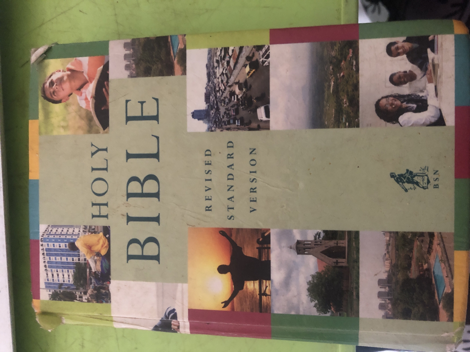 This media file displays the cover of a "HOLY BIBLE," specifically the "REVISED STANDARD VERSION," published by "BSN" (likely Bible Society of Nigeria), indicated by a logo of a figure possibly representing a sower or scholar. The cover features a collage of nine photographic panels arranged in a grid, set against a light green background with colorful borders.
**Subjects and Scenes:**
The panels depict a diverse range of subjects and settings, often blending spiritual, natural, and urban elements, likely reflecting life in locations like Abuja, Nigeria.
* One panel shows a young man with light skin, wearing a striped shirt, holding a book, and looking upwards with a serene expression, set in a sunlit outdoor, grassy area.
* Another features three young people – two males and one female with braided hair – smiling warmly at the camera, suggesting a group gathering or friendship.
* An aerial or high-angle shot captures a dense, bustling urban landscape with numerous buildings, cars on roads, and patches of green, indicative of a growing city like Abuja.
* A different urban scene displays a person in a yellow top and dark pants bending over near a tall, modern blue and white building.
* A striking panel presents the silhouette of a person with outstretched arms facing a vivid sunset or sunrise over a body of water, conveying a sense of worship or spiritual awe.
* Other panels include a traditional church building with a bell tower amidst trees under an overcast sky, and a modern urban skyline with high-rise buildings, green spaces, and a swimming pool, seen from a distance.
* Another panel shows a misty or cloudy landscape, and one features a partial view of a person wearing a striped arm sleeve, possibly hinting at activity or participation.
**Activity and Emotions:**
Activities include reading or contemplation, group interaction, urban life, and worship. Emotions conveyed range from serenity and joy to thoughtfulness and reverence.
**Notable Details:**
The time of day varies across panels, from bright daylight to sunset/sunrise. Weather conditions also differ, from clear skies to overcast. Location cues strongly suggest a vibrant, growing urban environment with both modern and traditional architectural elements, alongside natural beauty. The diversity of people and settings suggests a broad appeal and representation, characteristic of a global city. The collage format symbolically represents different facets of life and faith.