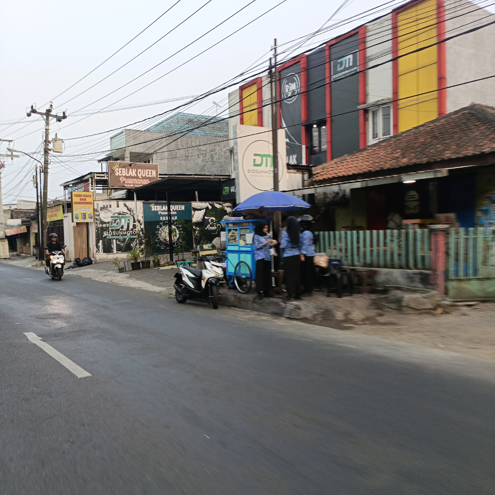 This image captures a street scene in Salatiga, Indonesia, likely in the afternoon given the lighting. The foreground shows a wet asphalt road with a white lane marking. On the right side of the road, there's a sidewalk with several establishments. Dominating the right side is a building with a prominent red and yellow facade, marked with black panels. A red tile roof is visible in front of this building. Next to it is a smaller structure with a teal-colored outdoor food cart selling what appears to be beverages or snacks, covered by a blue umbrella. Three women, dressed in blue shirts and black skirts or hijabs, are standing near the food cart. One is facing away, holding what looks like a paper cup, while the other two are facing the cart. A white motorcycle is parked to the left of the food cart. Further down the street, a man on a white scooter is riding towards the camera, wearing a black shirt and helmet. Above the buildings, numerous power lines crisscross the sky, supported by utility poles. Signs visible include "SEBLAK QUEEN Prasmanan" and "TOKO KACA DAN ALUMUNIUM," indicating local businesses. The overall atmosphere suggests a busy street with small businesses and everyday urban activity.