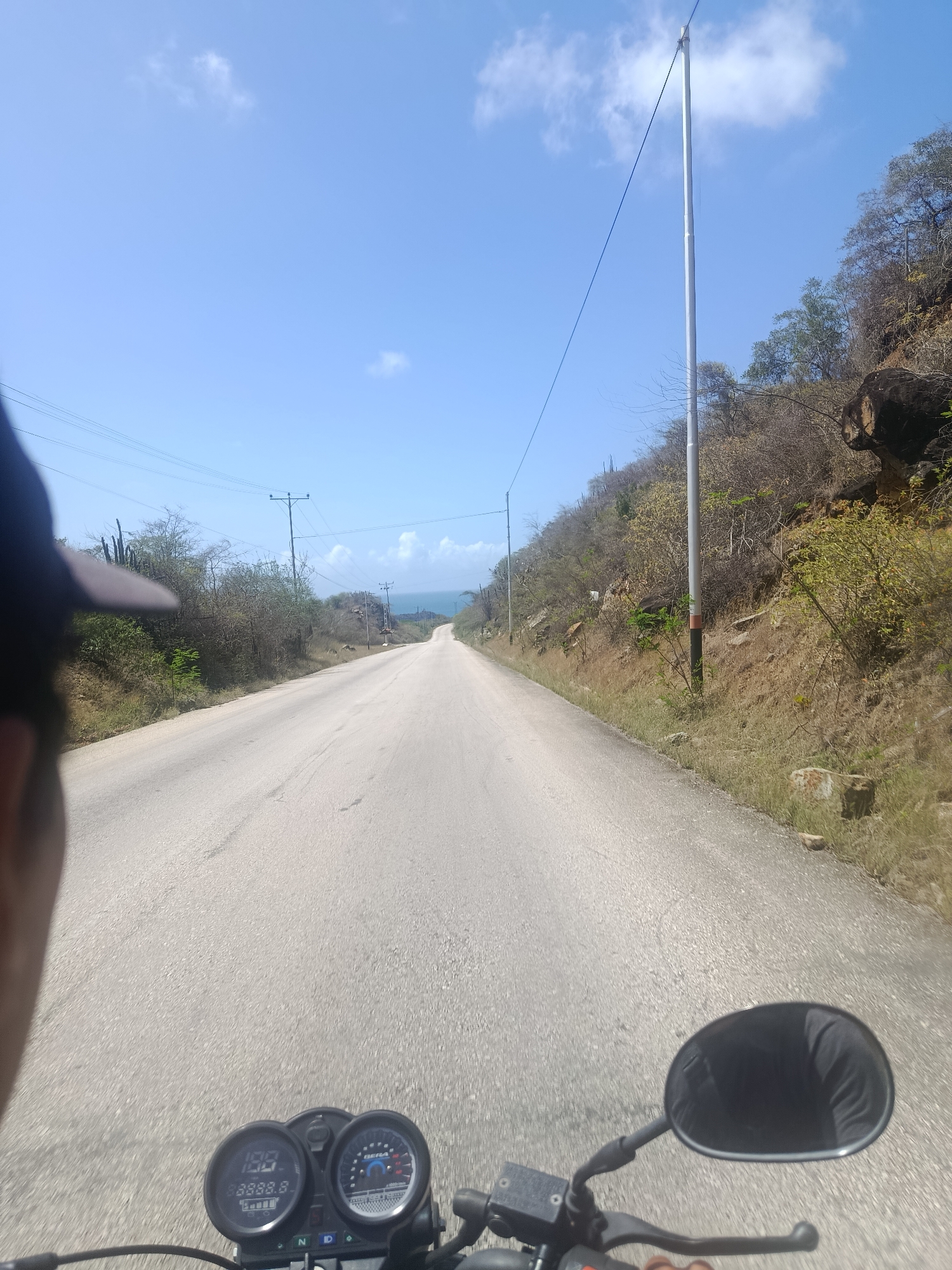 The image is a first-person perspective taken from a motorcycle on a paved road in Guayacán, Venezuela. The road stretches forward and curves slightly to the left, leading towards the horizon where the blue sky meets the blue ocean. The right side of the road is bordered by a dry, rocky hillside covered in sparse vegetation, including shrubs and some larger trees. Several utility poles with wires are visible along the roadside. In the foreground, the motorcycle's handlebars and a portion of the dashboard with a digital speedometer displaying "100" are visible. A black rearview mirror is also attached to the handlebars. The sky is clear blue with a few scattered white clouds. The overall scene depicts a sunny day on a rural road near the coast.