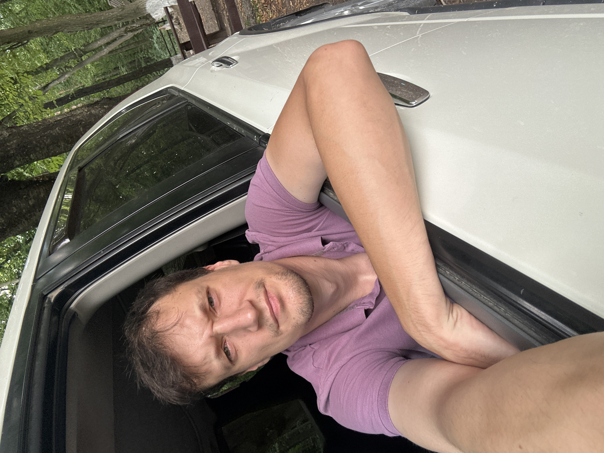 A man with short, brown, messy hair and stubble is leaning out of the open driver's side window of a white SUV. He is wearing a purple polo shirt. His arms are resting on the door frame. He is looking directly at the camera with a stern expression. The SUV is parked on a dirt path next to a wooded area with trees and some picnic tables visible in the background. The trees have green leaves. The lighting suggests it is daytime. The location is identified as Shatava, Ukraine.