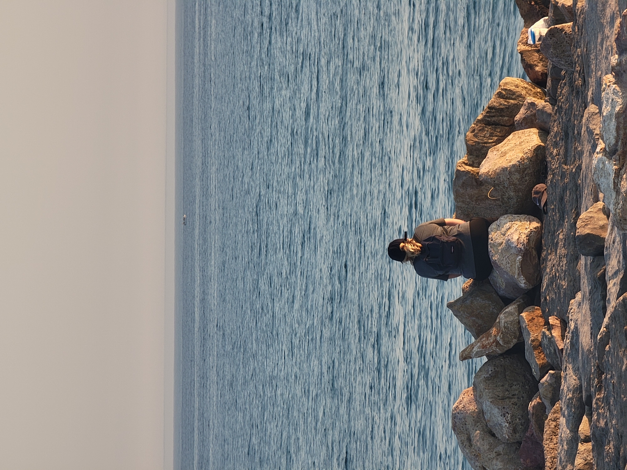 A person with a black baseball cap and blonde hair pulled back into a ponytail is sitting on a rocky shoreline, facing the ocean. They are wearing a grey t-shirt and a black backpack. The person is positioned in the center of the frame, with the ocean stretching out behind them and a faint horizon line visible in the distance. The foreground is composed of various-sized rocks and stones, some of which are damp. The water appears calm with gentle ripples. The sky is a pale, hazy color, suggesting either early morning or late afternoon light. A small white boat can be seen on the horizon in the distance. A white plastic bag is visible amongst the rocks on the right side of the frame. This scene is consistent with the natural rocky coastline found in various areas of Quarteira, Portugal, particularly along its expansive beaches and coastal paths.