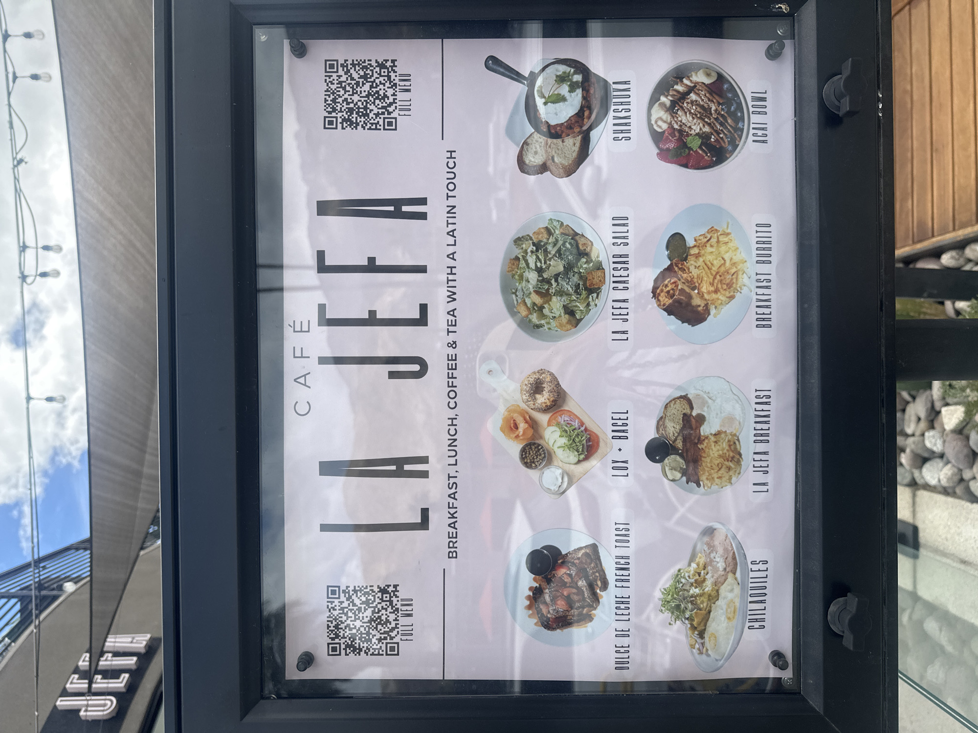 A close-up shot depicts a cafe menu displayed in a black frame. The menu lists various breakfast and lunch items. The text on the menu includes "CAFÉ LA JEFA" and "BREAKFAST, LUNCH, COFFEE & TEA WITH A LATIN TOUCH." Food items listed on the menu with accompanying images are Shakshuka, Acai Bowl, Breakfast Burrito, La Jefa Caesar Salad, La Jefa Breakfast, Lox Bagel, Dulce de Leche French Toast, and Chilaquiles. There are QR codes labelled "FULL MENU" in the top left and top right corners. The location is a cafe in the city of Unknown, Unknown. Outside the menu, the surrounding environment includes part of the cafe exterior, sky, and some landscaping.