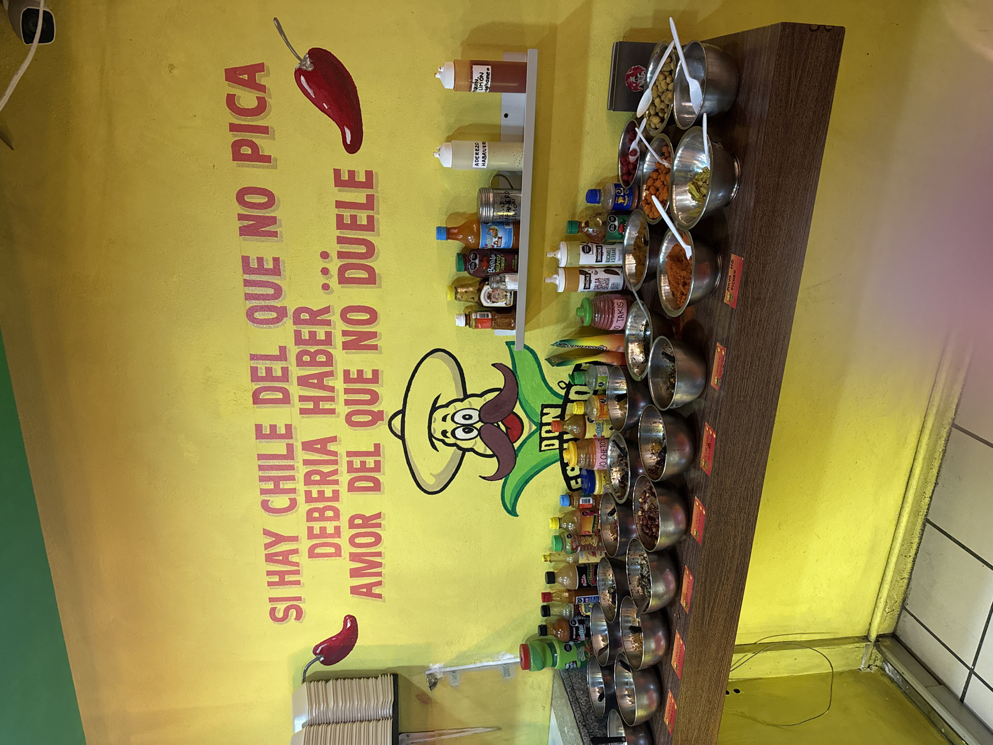 This image captures an indoor condiment bar within a food establishment located in San Bernardino Tlaxcalancingo, Mexico. The primary background is a vibrant yellow wall, accented by a narrow green band at the ceiling line. A large mural dominates the wall, featuring red text that reads, "SI HAY CHILE DEL QUE NO PICA DEBERIA HABER... AMOR DEL QUE NO DUELE" (If there's chili that doesn't burn, there should be... love that doesn't hurt). This text is accompanied by two stylized red chili pepper illustrations and a cartoon character with a sombrero and mustache, partially labeled "DON". A white floating shelf on the wall holds four bottles, two of which are white squeeze bottles labeled "ADEREZO HABANERO" and "TAQUIN LIMON". A security camera is visible in the upper right corner of the wall.
A dark brown wooden counter, functioning as a topping station, spans the lower half of the image. It is densely populated with approximately twenty small, round, silver-colored metal bowls, each containing various dry condiments or food toppings such as nuts, seeds, dried fruits, spice powders, and olives. Many bowls are equipped with white plastic serving spoons. Behind these bowls, an assortment of smaller bottles containing sauces and seasonings is arranged directly on the counter. Small, red-bordered rectangular paper labels are placed beneath some bowls, indicating their contents, such as "POLVO NO PICOSO" (non-spicy powder). To the far left, a stack of white square paper products is present. The floor is composed of white square tiles with dark grey grout lines. No individuals are visible in the scene.