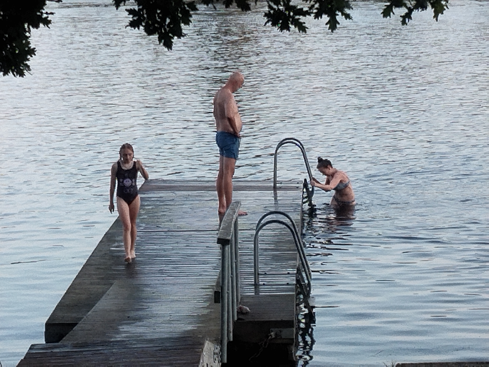 A full-body, low-angle shot depicts three individuals on a wooden pier and in the water. On the left, a young female with braided brown hair, wearing a black swimsuit with a pattern, walks barefoot on the pier away from the viewer. In the center, a bald man with a tan torso and wearing blue swim shorts stands facing away from the viewer, looking down. To the right, a woman with dark hair tied up, wearing a patterned bikini, holds onto the railing of a metal ladder as she partially submerges herself in the water. The water is a calm, blue-gray, with gentle ripples. The pier is made of dark, weathered wood planks. The background consists of more water, with branches of a tree visible in the upper left corner, suggesting an outdoor, possibly natural setting. The overall scene suggests a recreational activity, likely swimming, on a sunny day.