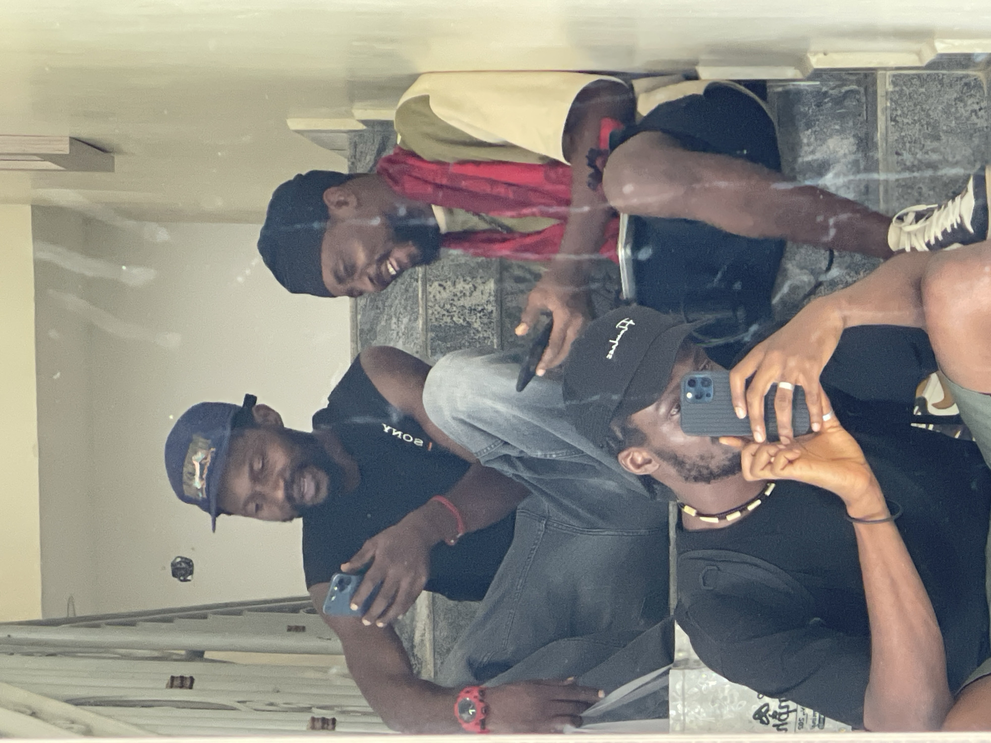 This vertical image is a reflection, likely taken in a mirror or large reflective surface, showing three young Black men posing together. The setting appears to be indoors, potentially within a building in Gwarinpa, Nigeria, indicated by the tiled steps or floor made of dark and light grey square tiles.
On the far left, a man wearing a dark baseball cap with faint graphics and a black t-shirt is visible, holding a blue smartphone in his left hand. He has a slight smile.
In the center, another man is seated, sporting a black beanie or durag, a light-colored top over a black t-shirt that displays the reversed word "SONY" across the chest, and grey jeans. A red scarf or fabric is draped around his neck. He is captured with a wide, joyful smile. He holds a dark smartphone in his right hand, and a red watch is prominent on his left wrist.
To the right, the third man is also seated, wearing a black baseball cap with some reversed text (partially visible). He is dressed in a black t-shirt and appears to be wearing black shorts. He is actively holding up a blue iPhone with multiple cameras, taking the reflection photo. He wears a beaded necklace and has white rings on his right hand.
The overall atmosphere conveyed is casual and friendly, with at least two of the men showing visible smiles, suggesting a cheerful gathering or group activity. The lighting is even, not indicating a specific time of day beyond being generally bright.