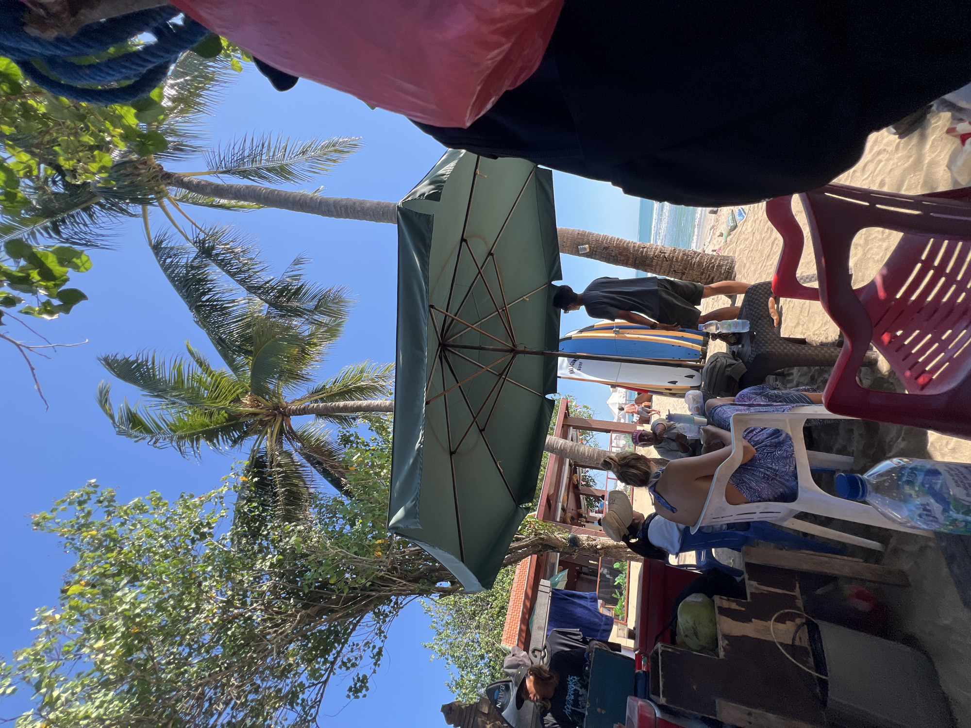 The image depicts a beach scene in Kuta, Indonesia, on a sunny day. In the foreground, a large green umbrella provides shade over a table where several people are seated. Two women are visible, one wearing a sun hat and the other with her back to the camera, seated on white chairs. A man wearing a dark t-shirt and shorts stands near surfboards. Behind them, palm trees and other vegetation are visible against a clear blue sky. Further in the background, the ocean waves can be seen, with a few people on the beach. The scene appears to be set up for beachgoers, with surfboards suggesting a surf-friendly location.
