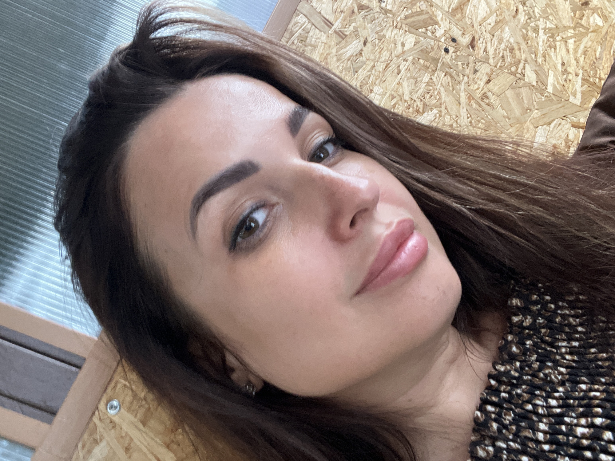 A medium shot shows a woman with dark brown hair from the chest up. She is looking directly into the camera, and her head is tilted to the left. The woman is wearing a black and tan patterned top. She has dark eyebrows, brown eyes, and a subtle smile. She is wearing makeup, including eyeliner and lipstick. She has a small stud earring in her visible ear. The background appears to be indoors and consists of light-colored wood panels and a gray corrugated surface above. There is a visible screw in the wood paneling behind her.