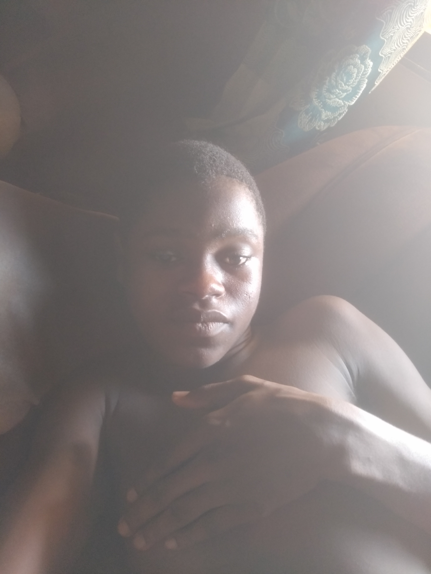 The image features a young person with dark skin and short, dark hair, possibly a woman, looking directly at the camera in what appears to be a selfie. They are bare-chested or wearing a very sheer, light-colored garment, with their right hand gently placed across their chest, covering part of their torso. Their left arm is also visible, resting near their side. The person has a neutral expression, with highlights visible on their forehead, nose, and lips due to strong lighting.
The scene is characterized by heavy overexposure from a bright light source, likely daylight, entering from the upper right. This creates a significant glare and haze that washes out much of the right side and upper portions of the image.
In the upper background, a dark fabric with a distinct, light-colored woven or embroidered floral pattern, resembling a rose, is visible. This suggests an indoor setting, possibly a curtain, a cushion, or part of a headboard. The remainder of the background is dark and indistinct. There is no visible text in the image. The context provided is Shangisha, Nigeria.
