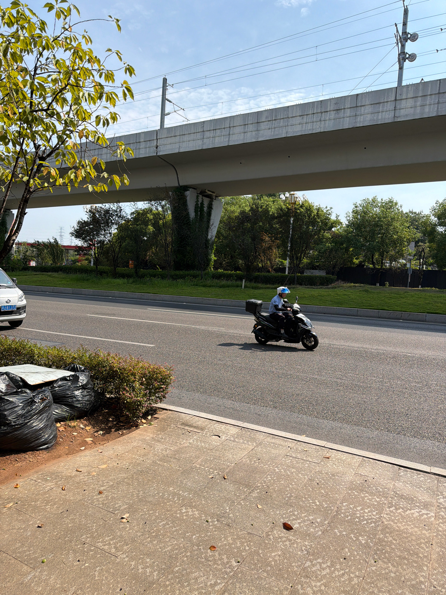 A person wearing a light blue helmet and a blue shirt is riding a black scooter with a black storage box on the back, on a paved road in the city of Jiangjin, China. The scooter is moving from left to right. In the foreground, there are several black trash bags piled up next to a row of green bushes. A concrete sidewalk with scattered leaves is visible to the left of the trash bags. In the background, an elevated concrete structure, likely a bridge or overpass for a railway, spans across the scene. Electrical wires are suspended above the elevated structure, supported by utility poles. Trees and green grass line the side of the road beyond the scooter. A white car with a license plate is parked on the left side of the road. The sky is clear with some scattered clouds.