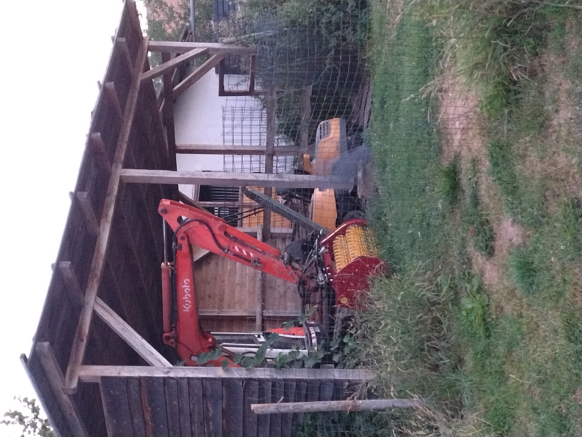 The image shows a red Kubota excavator parked under a wooden shelter in Dobev, Czechia. The shelter has a dark, planked wall on the left and a lighter-colored building behind it. The excavator is facing towards the right, with its arm extended and bucket down. The bucket appears to be a rototiller attachment with yellow tines. There is a wire fence in front of the excavator and shelter, with tall grass and weeds growing around it. Another yellow machine, possibly a small tractor or utility vehicle, is partially visible behind the excavator. The ground is covered in grass and some bare patches. The sky is overcast and the lighting suggests it is either early morning or late afternoon. There are no people visible in the image.