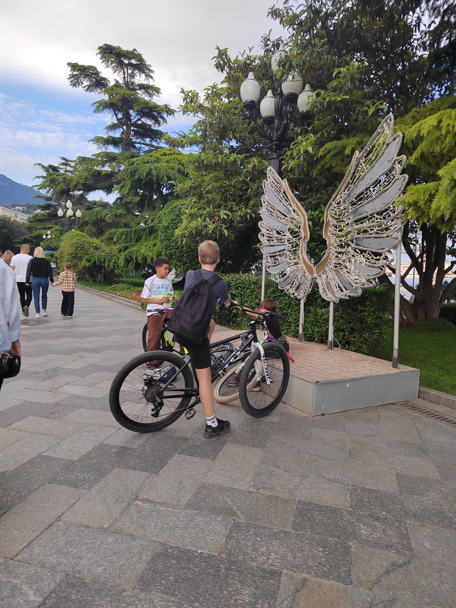 The image depicts a paved walkway with several people present. Two boys are standing with bicycles near a large metal sculpture of wings. Behind the boys and the sculpture, there's lush greenery, including trees and bushes. In the background, other people are walking along the path. There is a distinctive tree that is shaped like a candelabra. The area appears to be a park or pedestrian zone, likely in an urban setting with a visible street lamp nearby.
