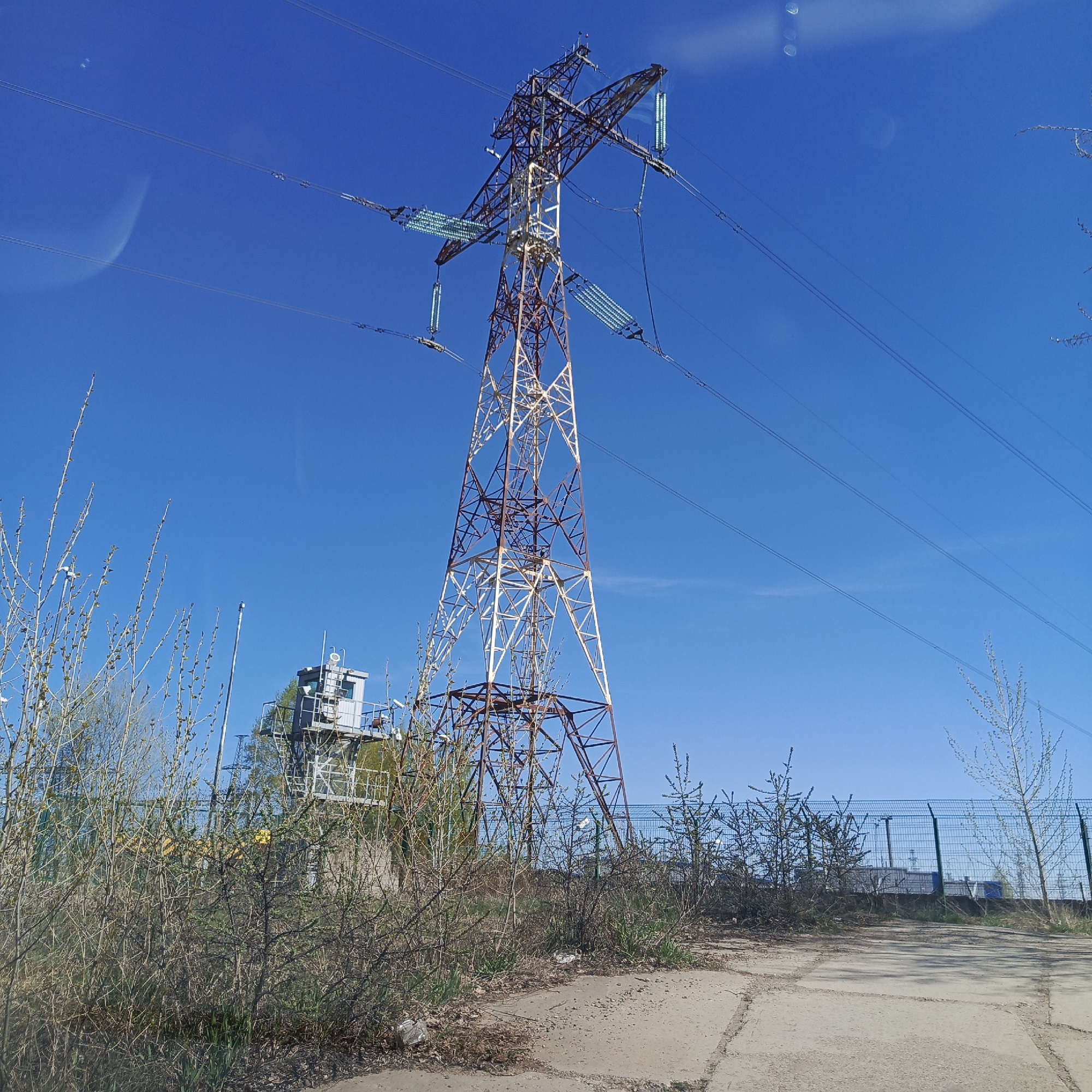 The image shows an electrical substation in Chaikovsky, Russia. A tall, lattice-style transmission tower dominates the scene. It is painted white and brown. High-voltage power lines extend from the tower against a clear blue sky. In the foreground, there is vegetation and a concrete area with some debris. A small control building is present to the left of the tower, inside a fenced area. There is a chain link fence in the background.
