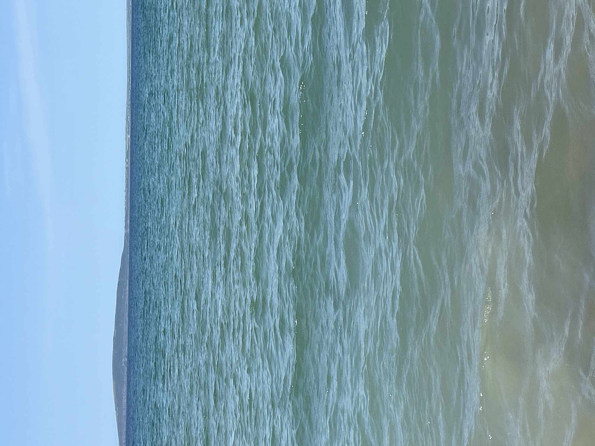 The image shows a wide expanse of calm, turquoise ocean water under a clear blue sky. In the distance, a low-lying island or peninsula with a dark, undulating silhouette is visible on the horizon, separating the water from the sky. The foreground of the image displays the gently lapping waves at the edge of the water, with the sandy seabed visible beneath the surface. The scene evokes a sense of tranquility and natural beauty. This location is consistent with the coastal landscape of Ballybunnion, Ireland, known for its scenic beaches and ocean views. There are no people or specific structures visible in the frame, emphasizing the natural environment.