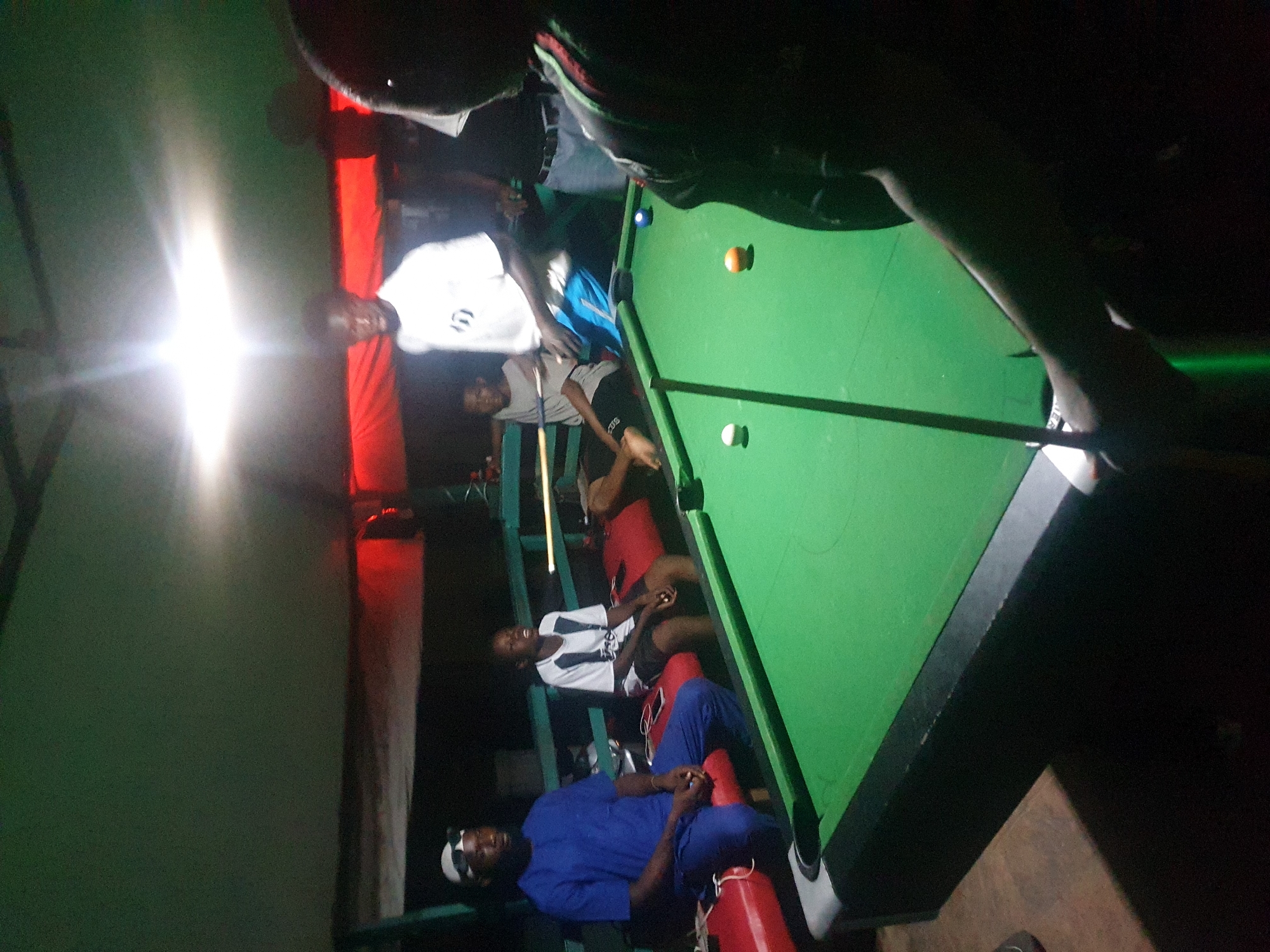 An undated, slightly blurry, medium shot photograph taken at night depicts a group of individuals engaged in a game of billiards within an indoor, dimly lit establishment. The setting appears to be a community or recreational space in Lokoja, Nigeria, suggested by the context.
In the foreground, the vibrant green felt surface of a pool table dominates the lower portion of the image. Several billiard balls are visible: a white cue ball, an orange solid ball, and a blue ball. A pool cue extends from the right side of the frame, held by an unseen person.
To the left of the pool table, two men are seated on a red bench. The man closest to the camera wears a blue traditional garment and a white head covering with sunglasses. He has a serious expression and his hands are clasped in his lap. Next to him, a younger individual in a white and black t-shirt and dark shorts is also seated, looking towards the pool table with his mouth slightly open.
Further back, a young man in a white t-shirt and blue shorts stands behind the pool table, his stance suggesting he might be playing. Another individual is partially visible behind him, also standing. The background features a dark wall with a red horizontal band across it, likely a decorative element or lighting feature. Overhead, a green tarp or ceiling is visible, illuminated by bright, harsh overhead lights that create a halo effect. The overall atmosphere is casual and convivial.